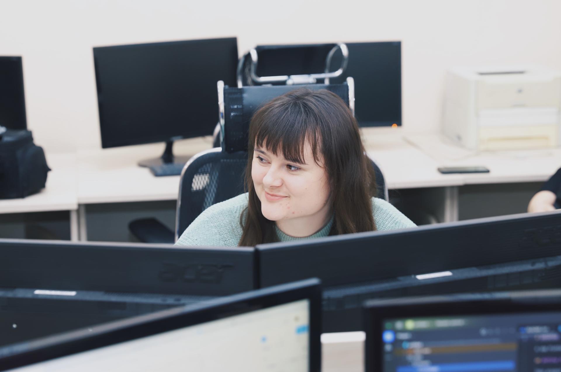 A young woman with long hair smiles while seated at a desk with multiple computer monitors.