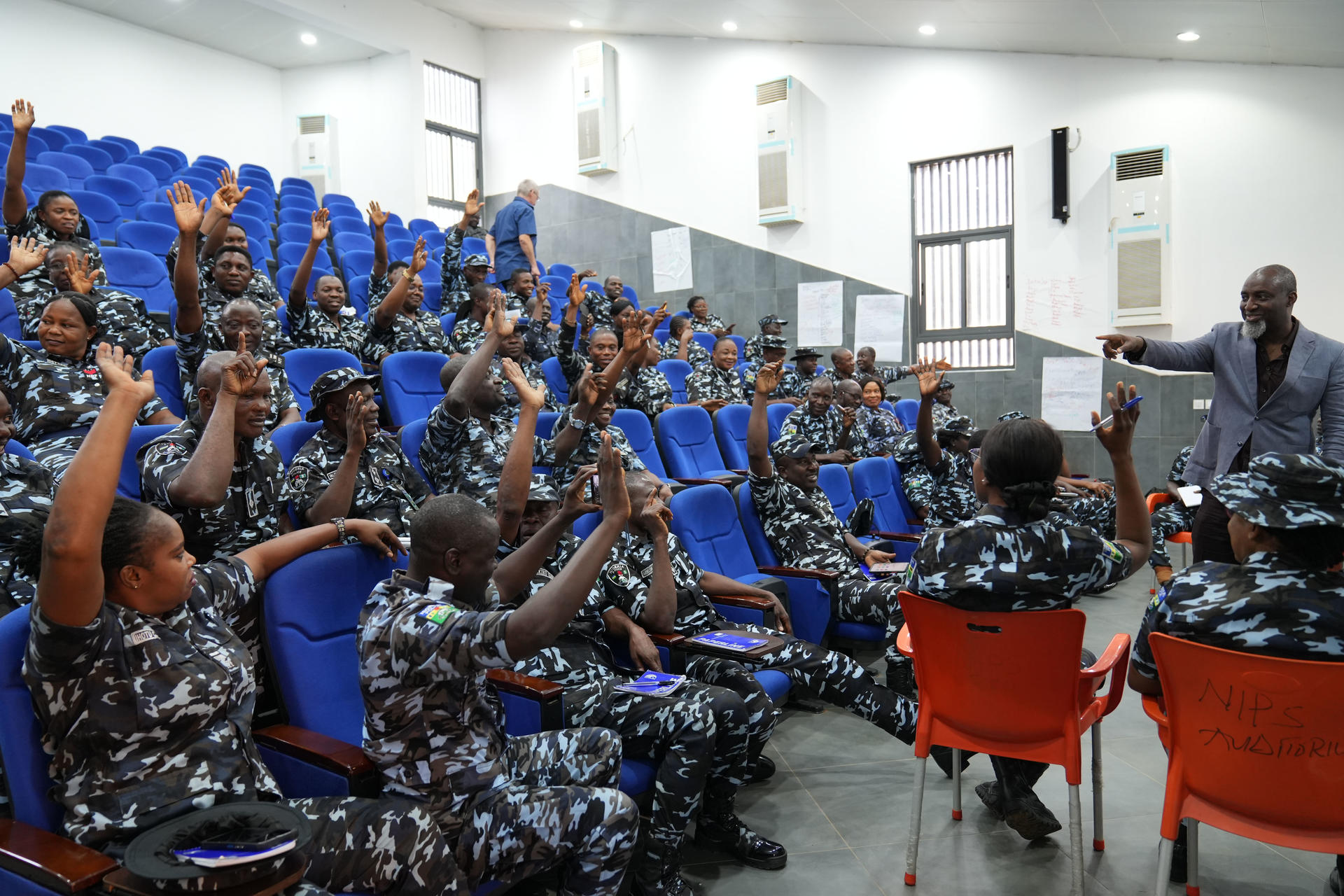 A group of uniformed individuals in a classroom raising their hands while a speaker gestures.