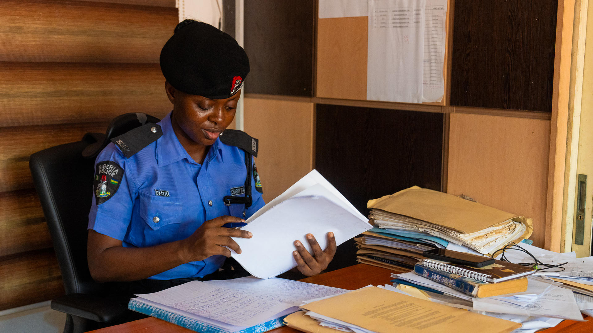 A police officer in uniform sitting at a desk, reviewing documents among stacks of paperwork.