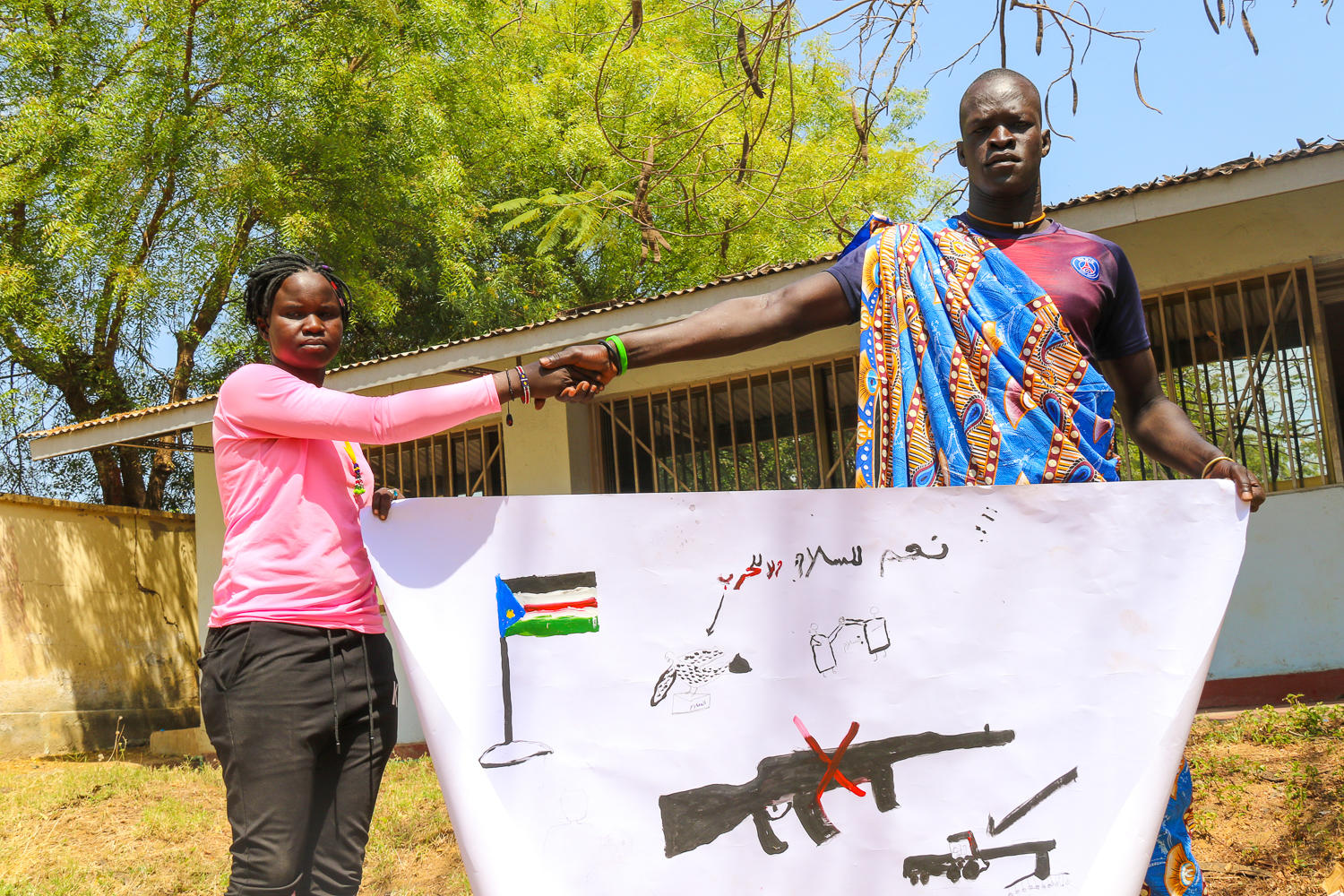 Two individuals shake hands while holding a large banner with symbols and a peace message.