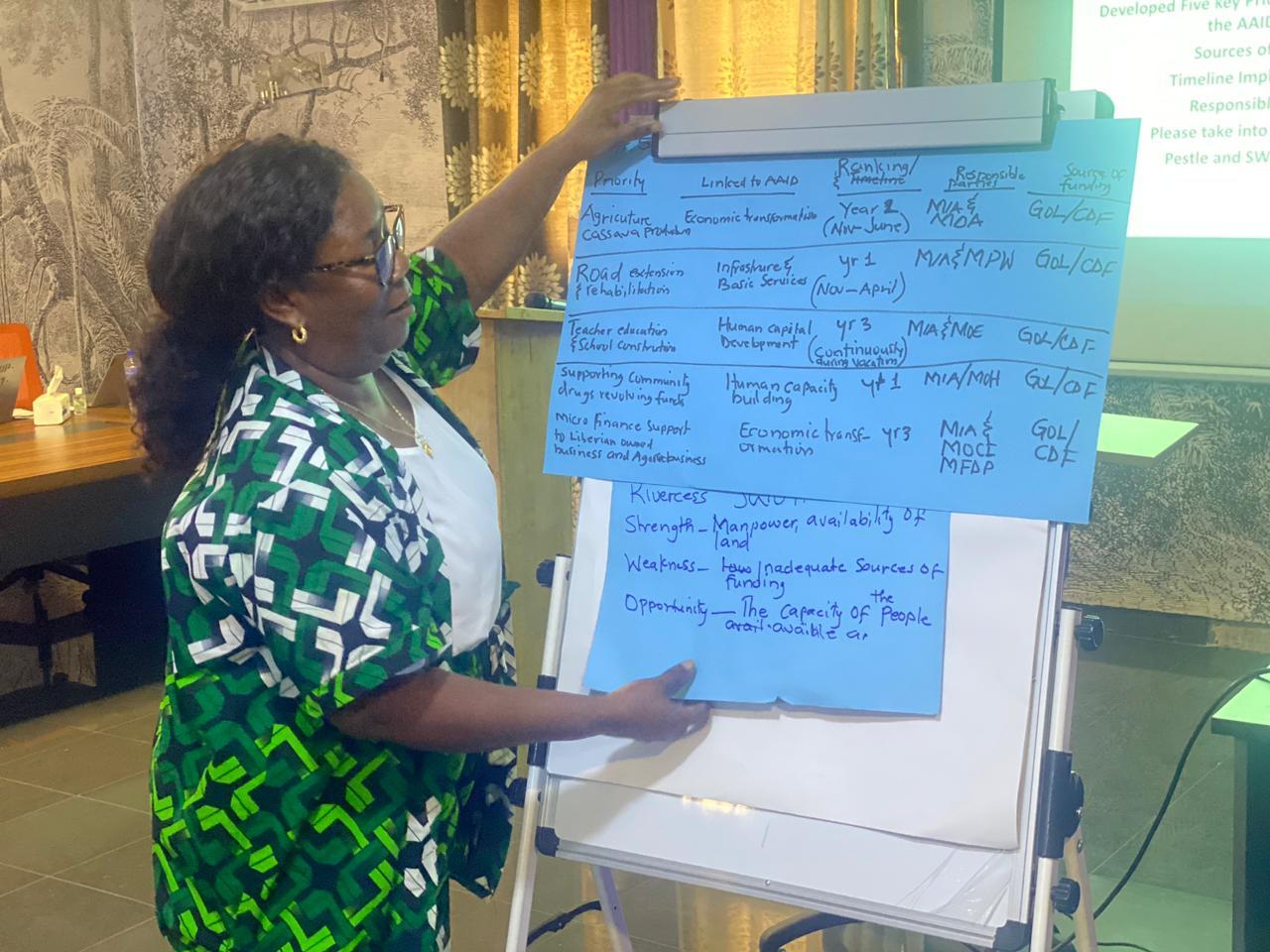 A woman in a patterned green and black outfit holds a blue chart on an easel in a meeting room.