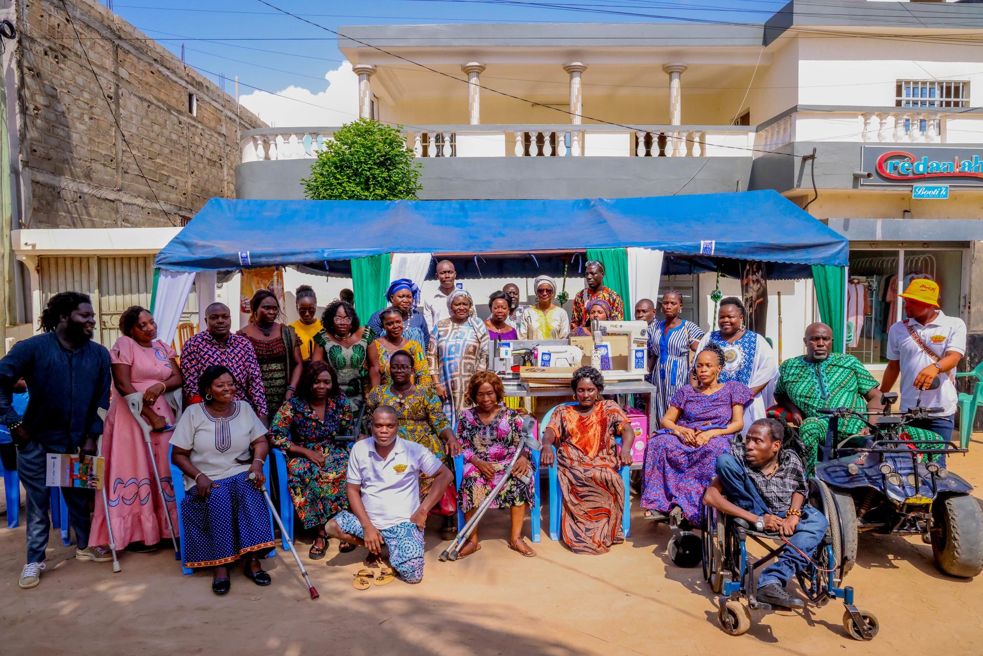 A diverse group of people posing together outdoors, some in wheelchairs, in colorful attire.