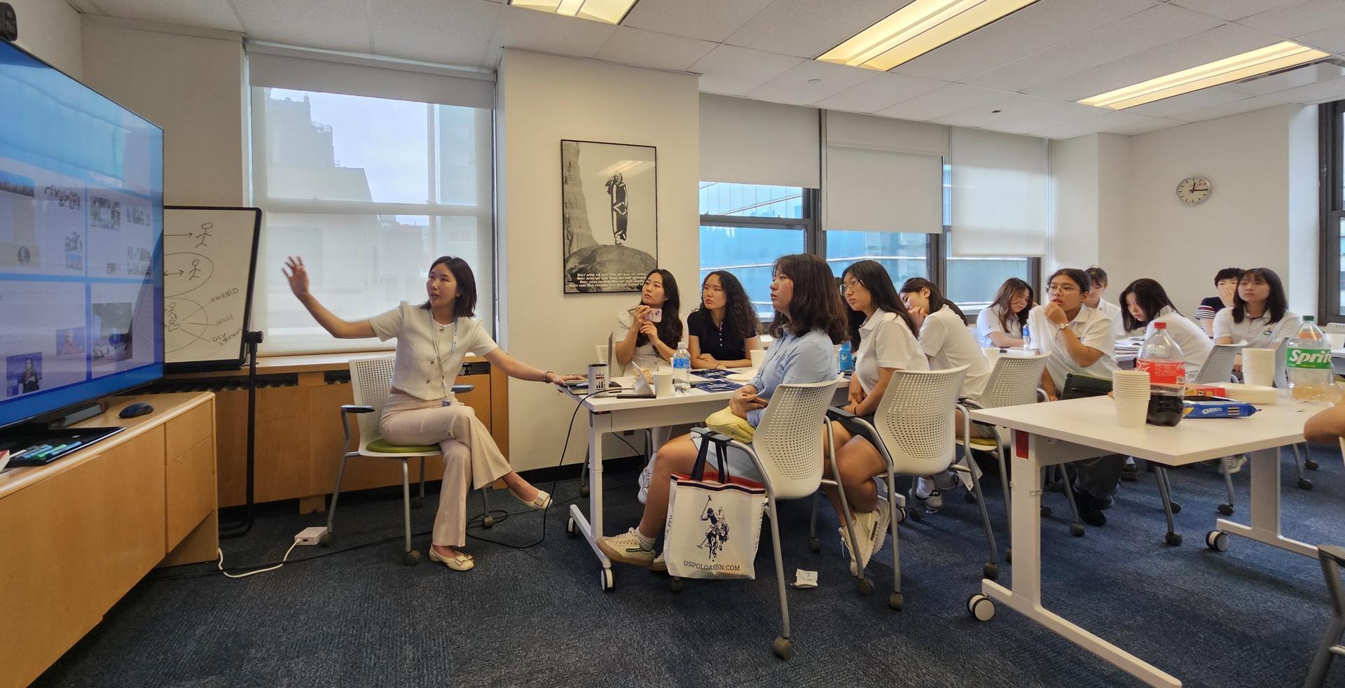 A teacher gesturing while leading a lesson to a group of attentive students in a classroom.