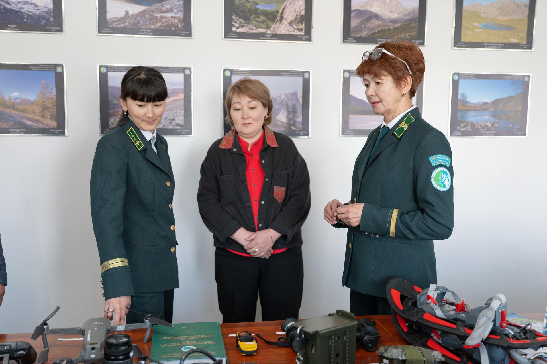 Three women in uniforms are examining equipment on a table with framed landscape photos in the background.