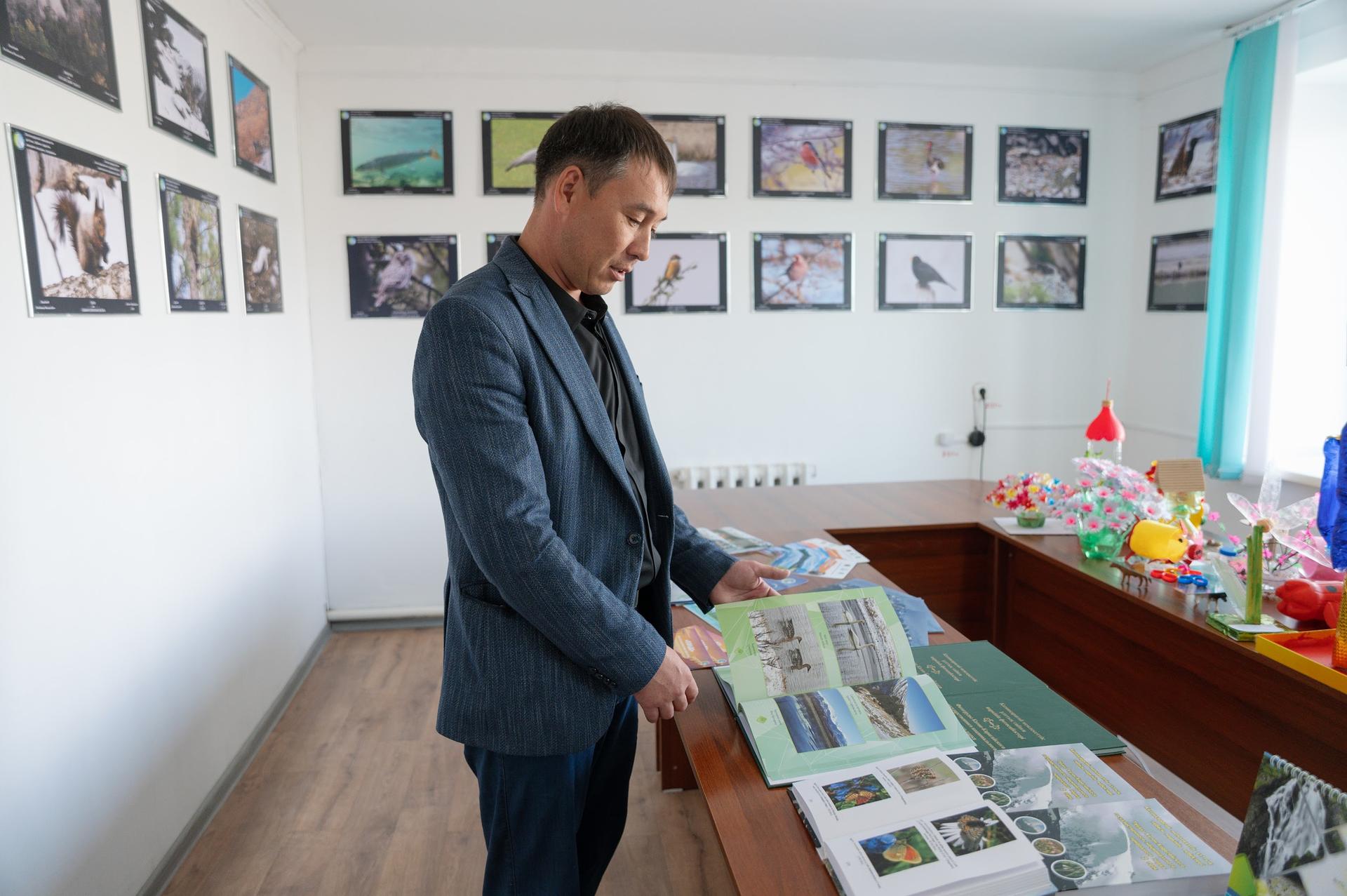 A man in a suit examines a book at a table in a room with framed photographs on the walls.