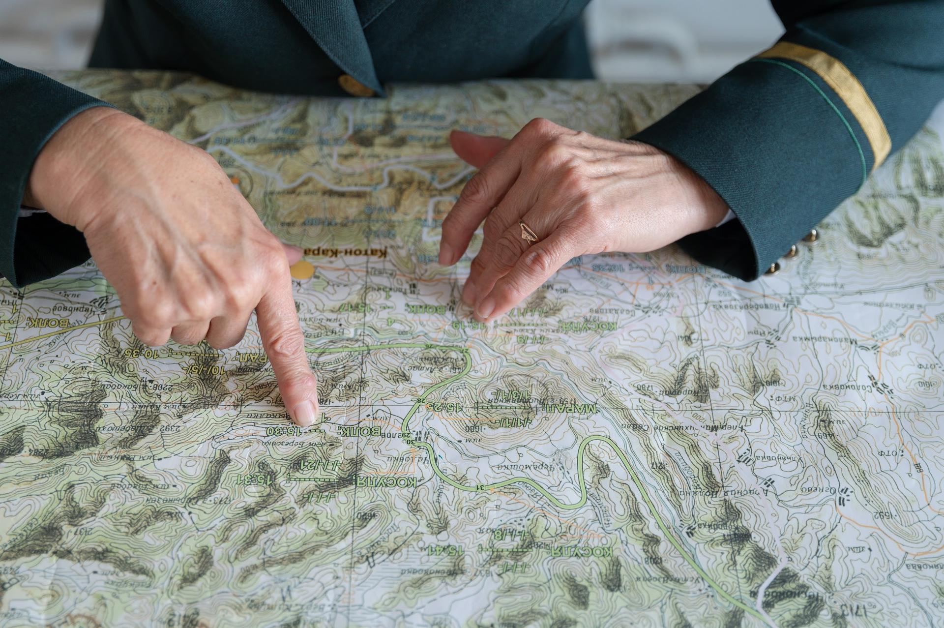 A person in a green uniform points at a detailed map spread on a table.