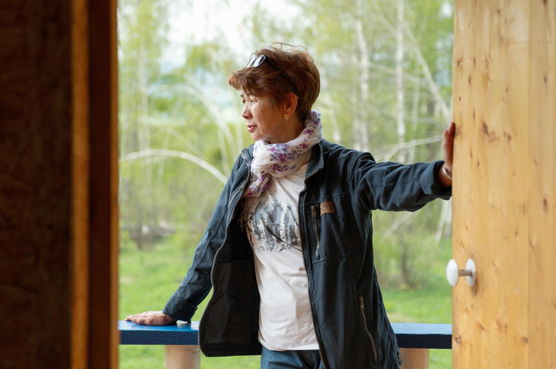 A woman with short hair stands by a wooden door, looking thoughtfully outside at greenery.