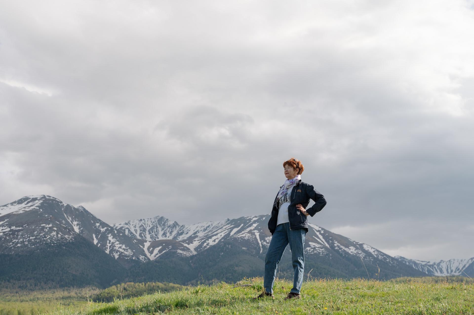 Person standing confidently on a grassy hill with snow-capped mountains in the background.
