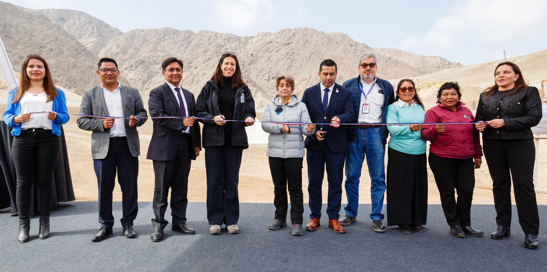 A group of eight people, dressed in formal attire, holding a ribbon outdoors in a mountainous area.