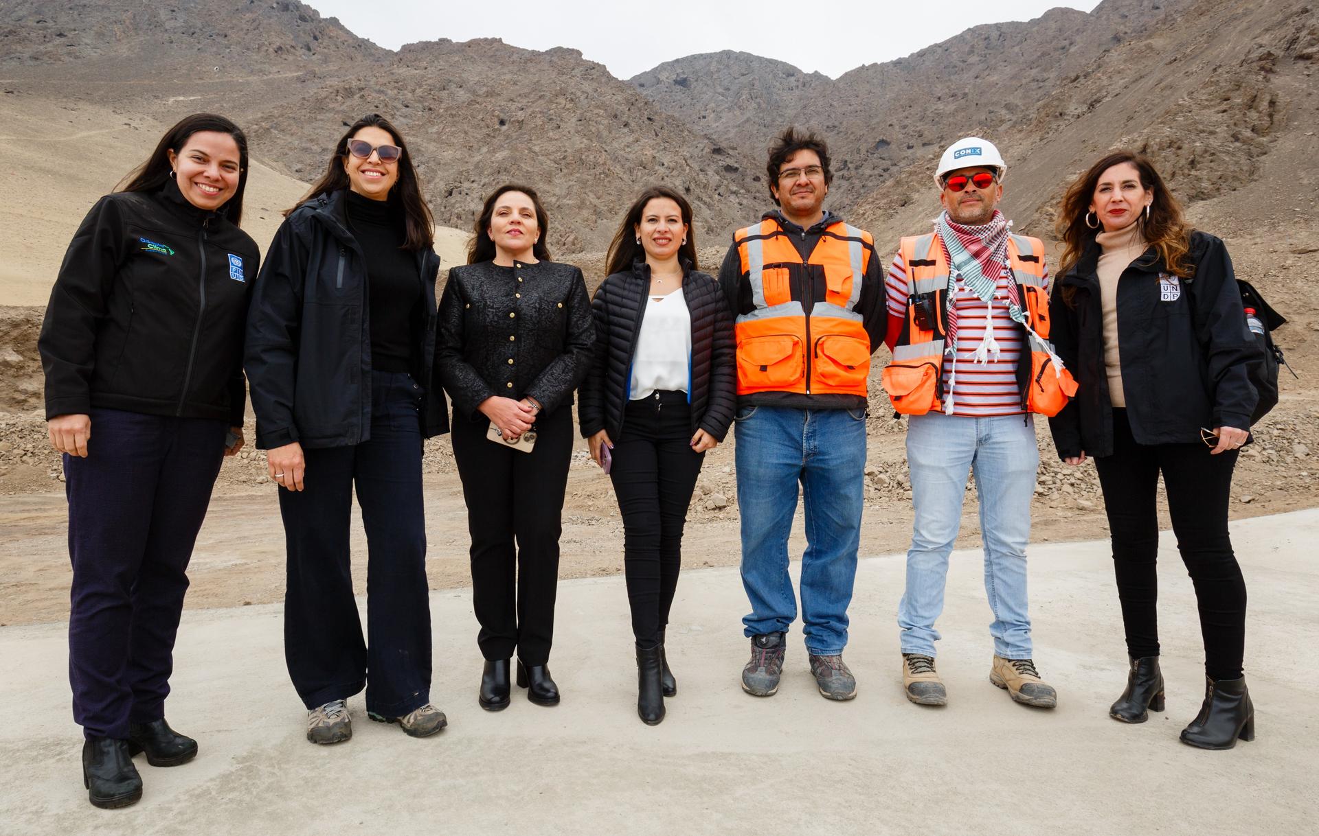 A group of seven people stand together outdoors in a mountainous landscape, smiling.