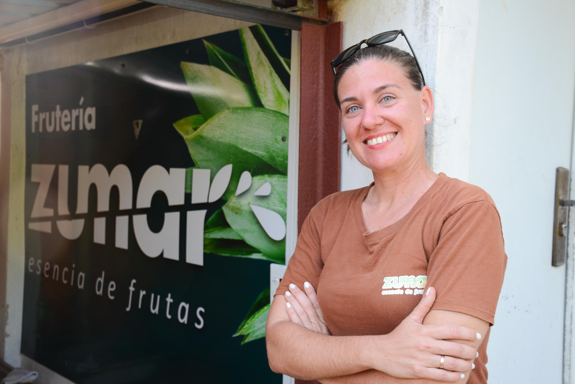 Smiling woman in a brown shirt stands confidently in front of a fruit store sign.