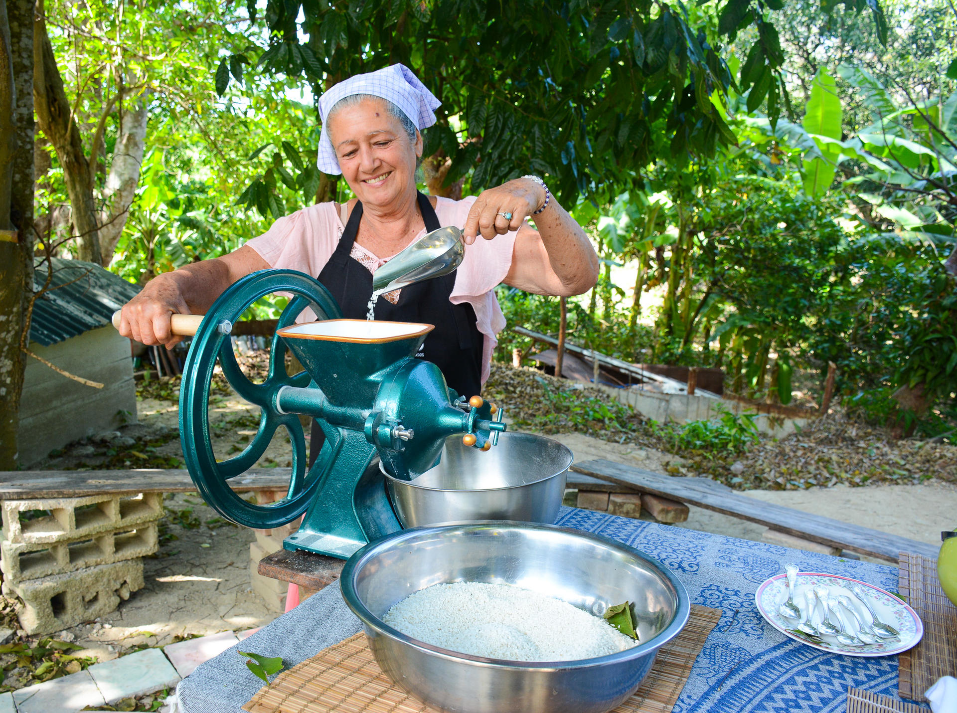 Woman using a traditional manual grinder outdoors, surrounded by greenery and bowls of food.