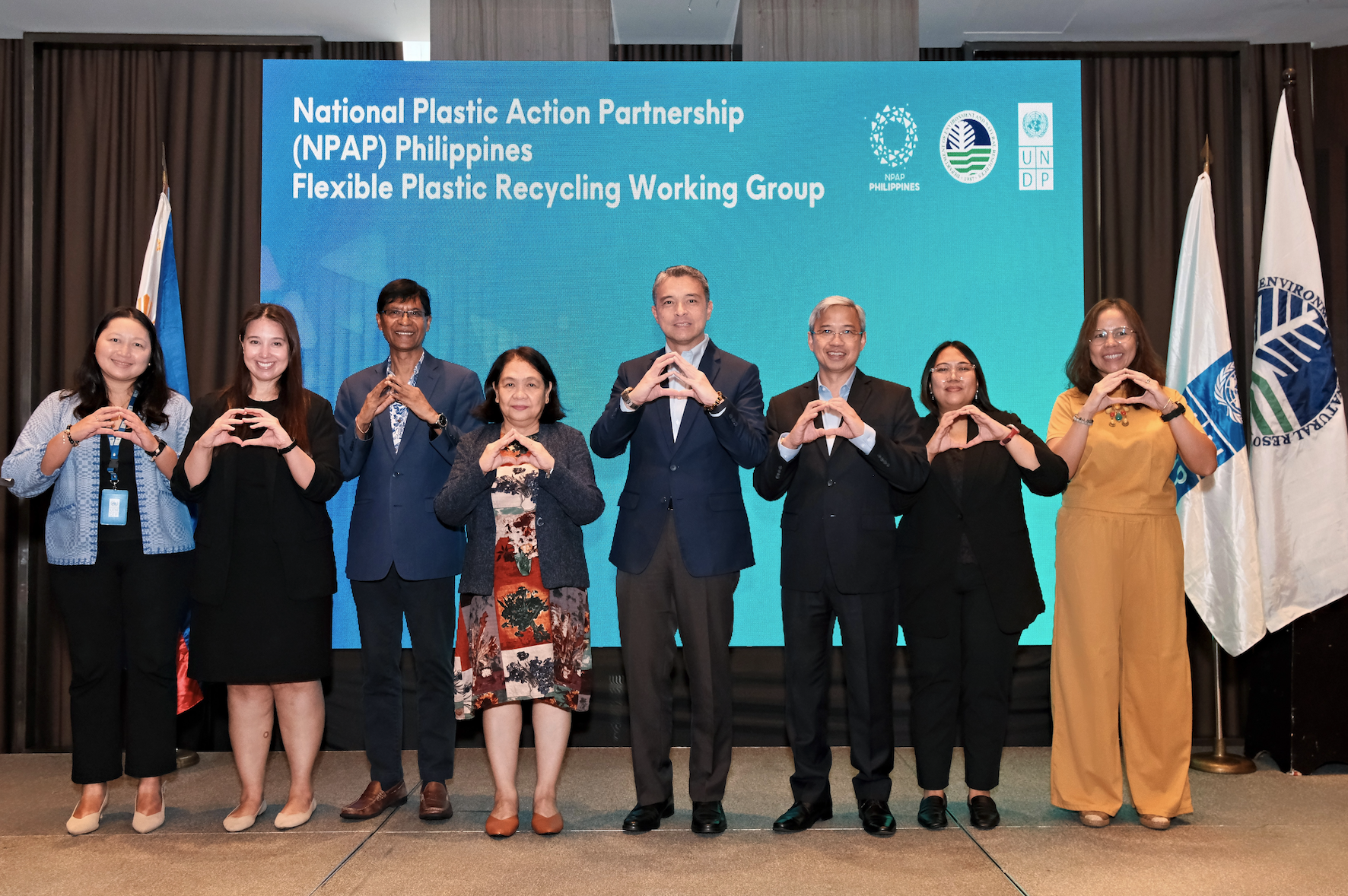 Eight individuals stand together, forming a heart shape with their hands in front of a conference backdrop.