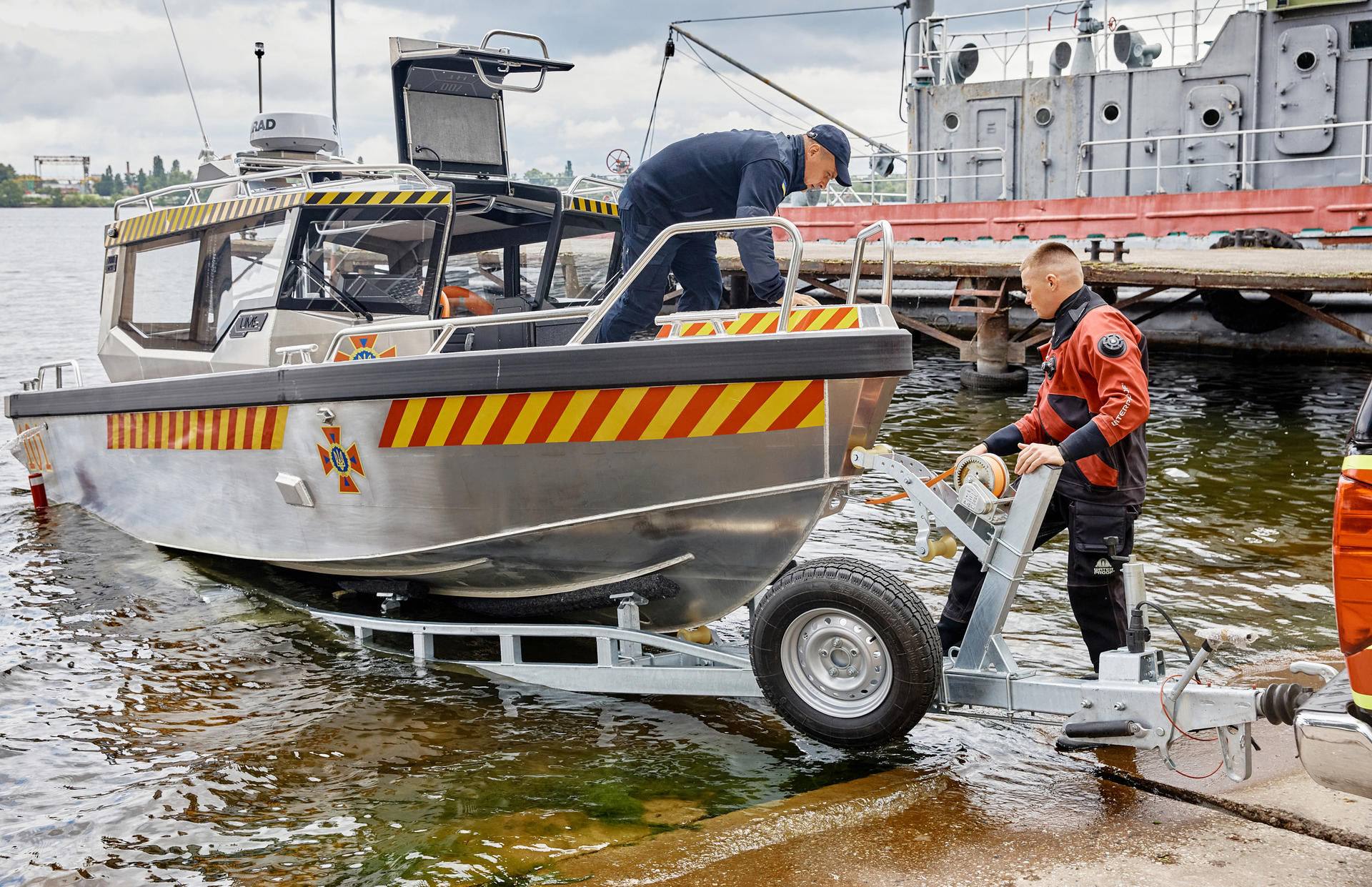 A small boat is being launched from a trailer by two individuals on the shore.