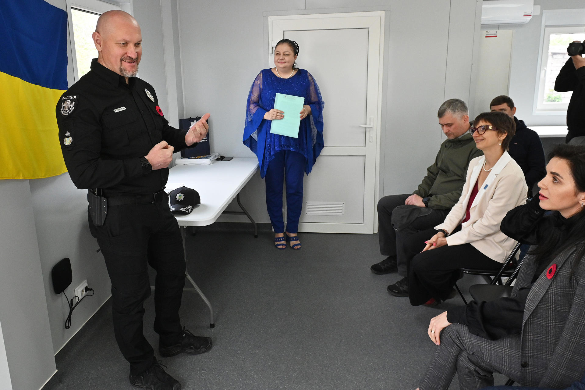 A police officer speaks to a group of seated individuals in a room with a Ukrainian flag.