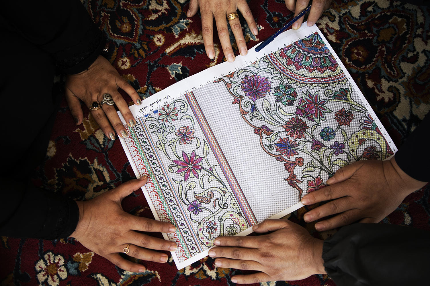 Hands gathered around a brightly colored design book on a patterned rug.