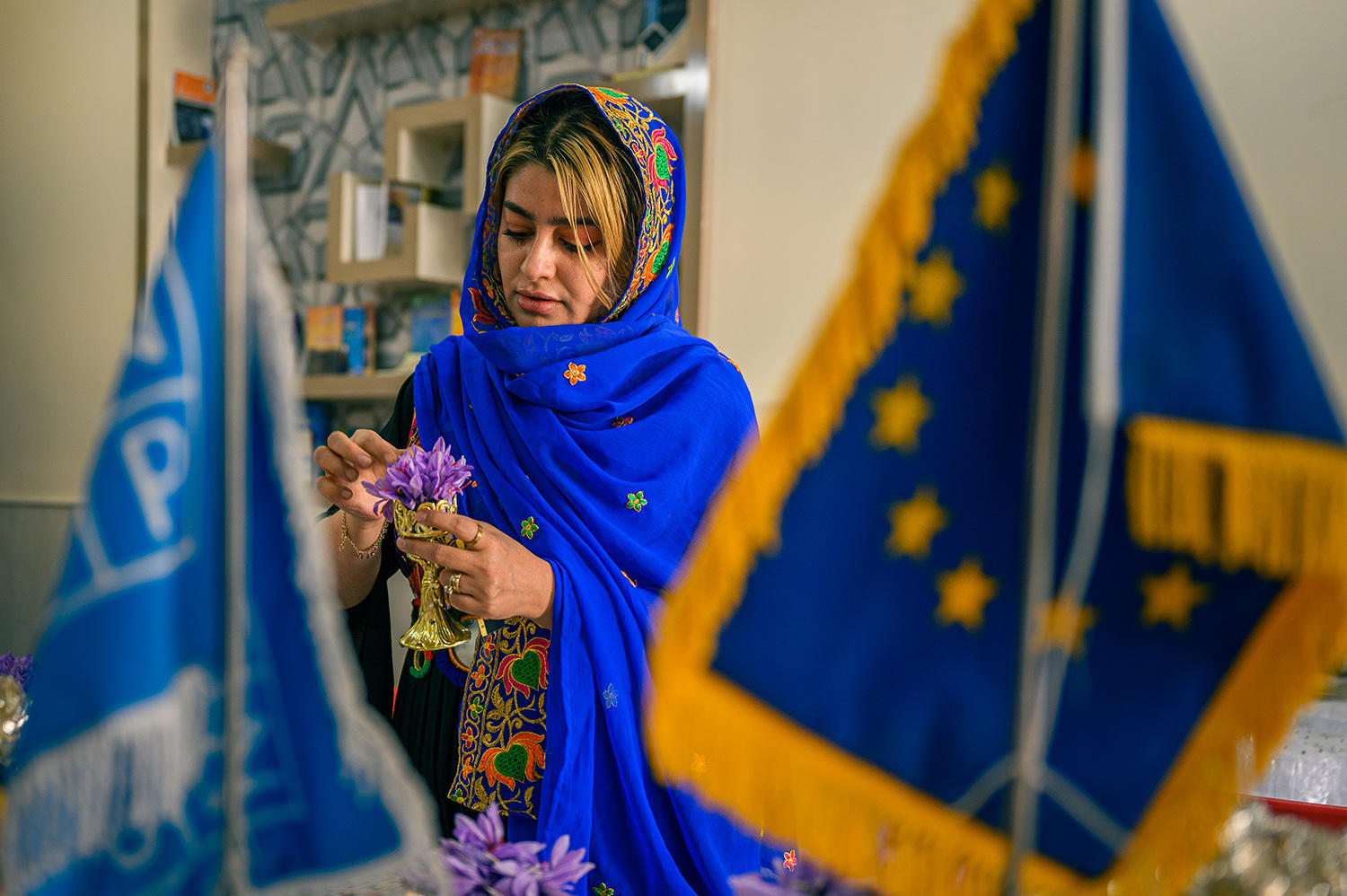 A woman in a blue shawl arranges flowers between flags, displaying cultural pride.