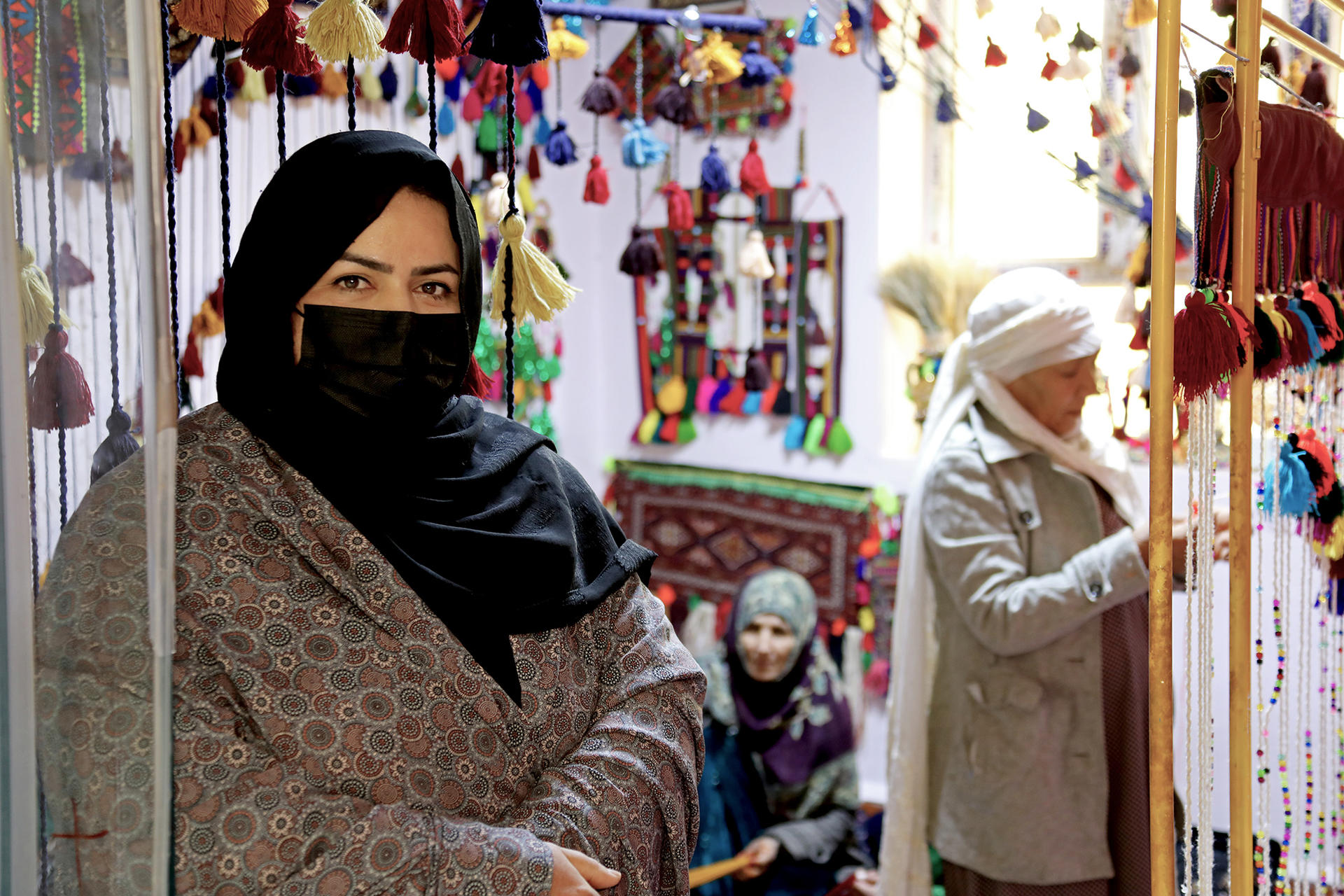 A woman in a black hijab stands in a vibrant shop, surrounded by colorful tassels and decorations.
