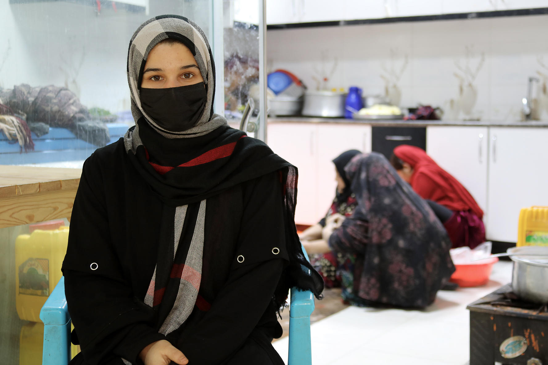 A woman in a black hijab and mask sits in a brightly lit kitchen while two others prepare food.