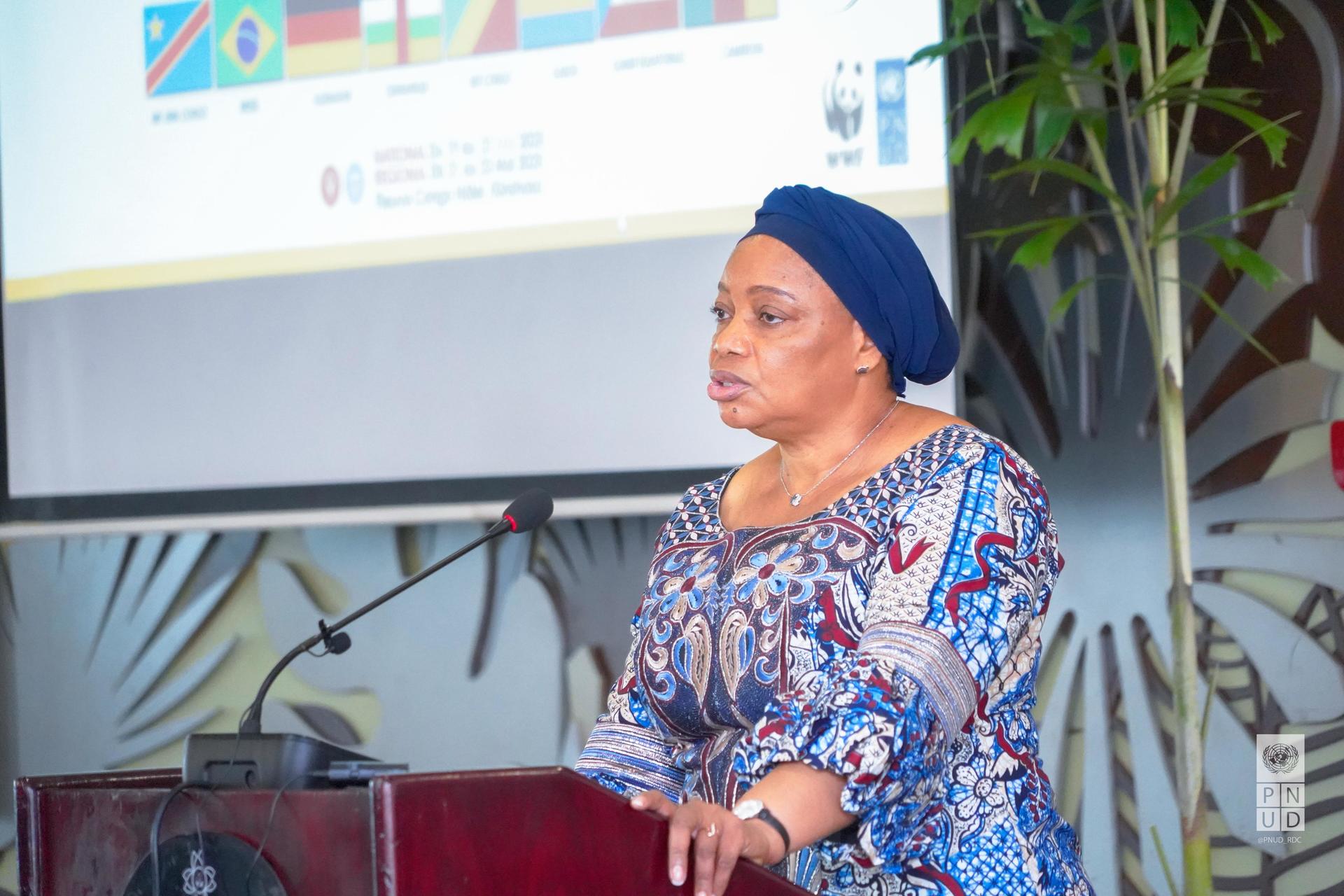 A woman wearing a patterned dress and headscarf speaks at a podium with a presentation behind her.