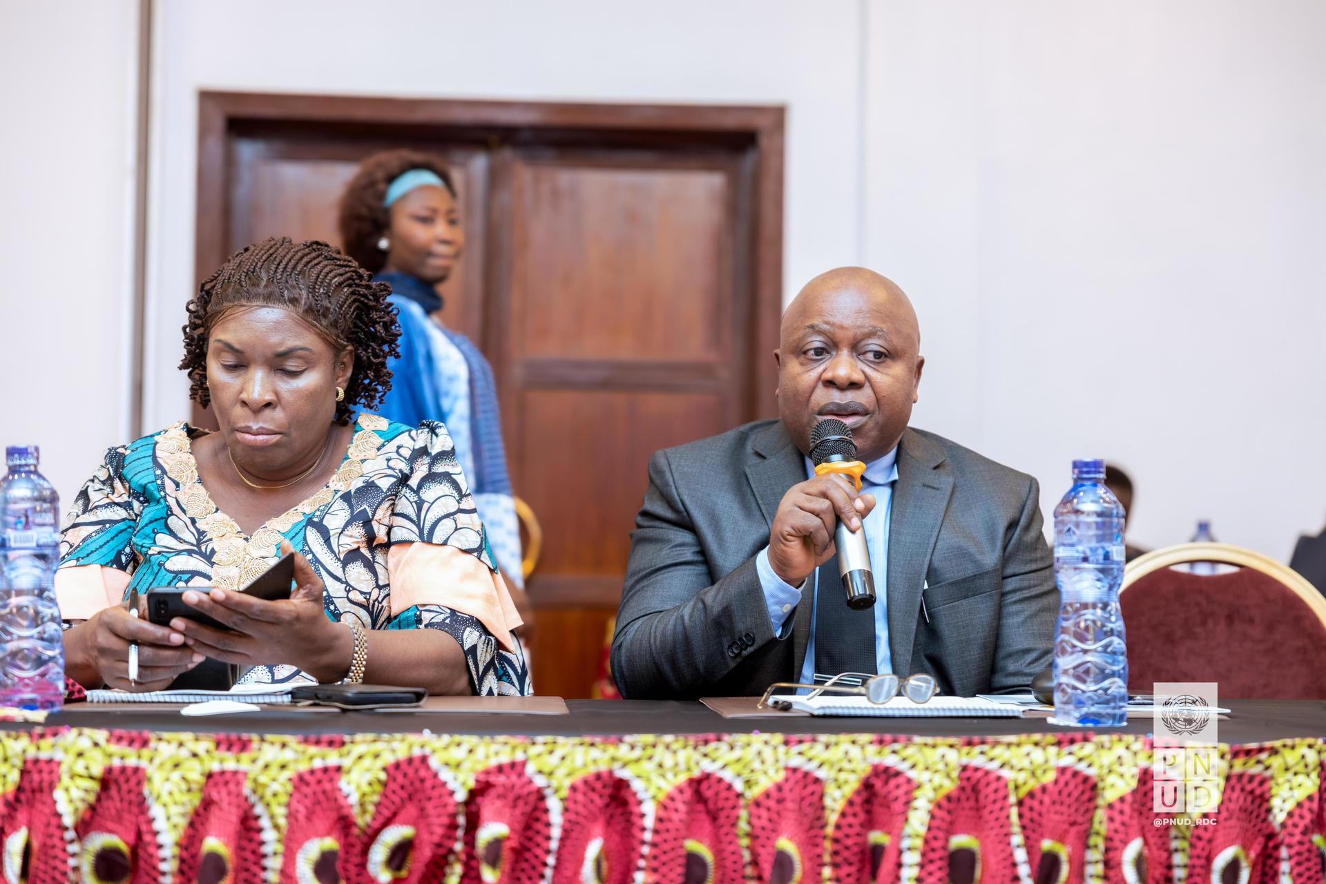 A man speaking into a microphone at a conference table, with a woman listening.