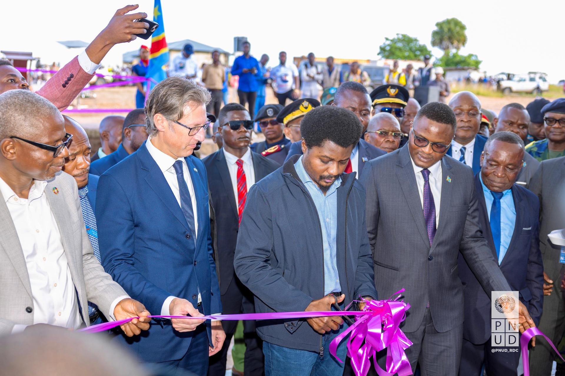 A group of officials gathered around, preparing to cut a purple ribbon at an event.
