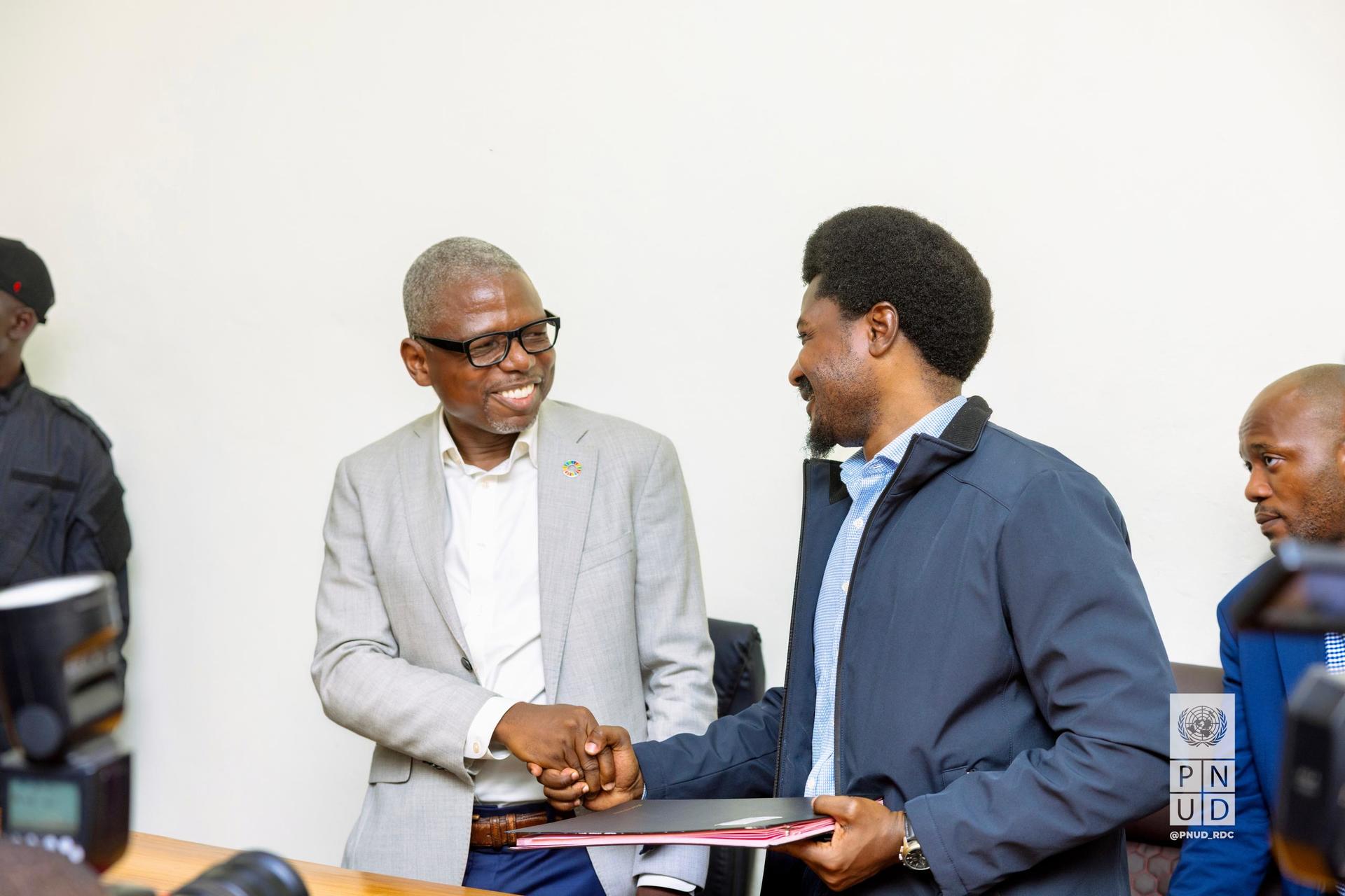 Two men shake hands and smile, surrounded by a group in a formal setting.