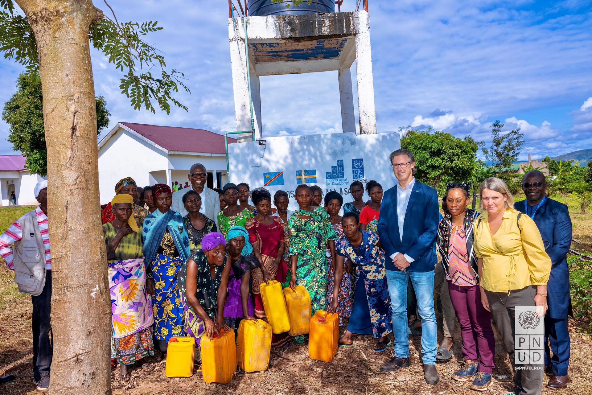 Group of men and women with water jugs, standing in front of a water tower and buildings.