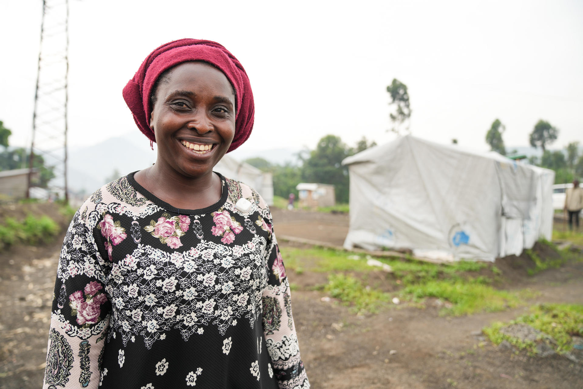 A smiling woman in a pink headwrap stands outdoors near a makeshift tent.