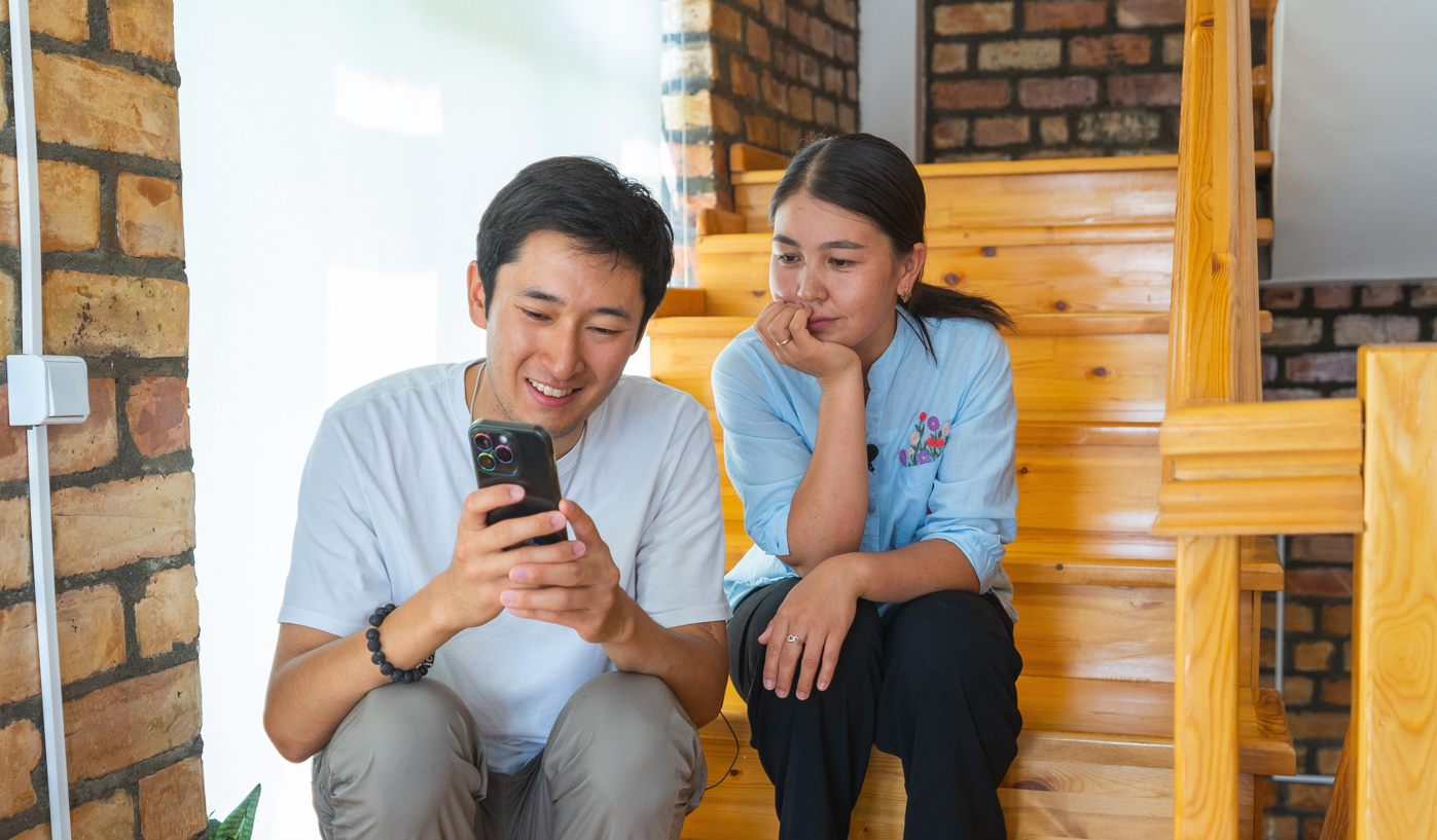 A young man and woman sit on wooden stairs, smiling at a smartphone together.