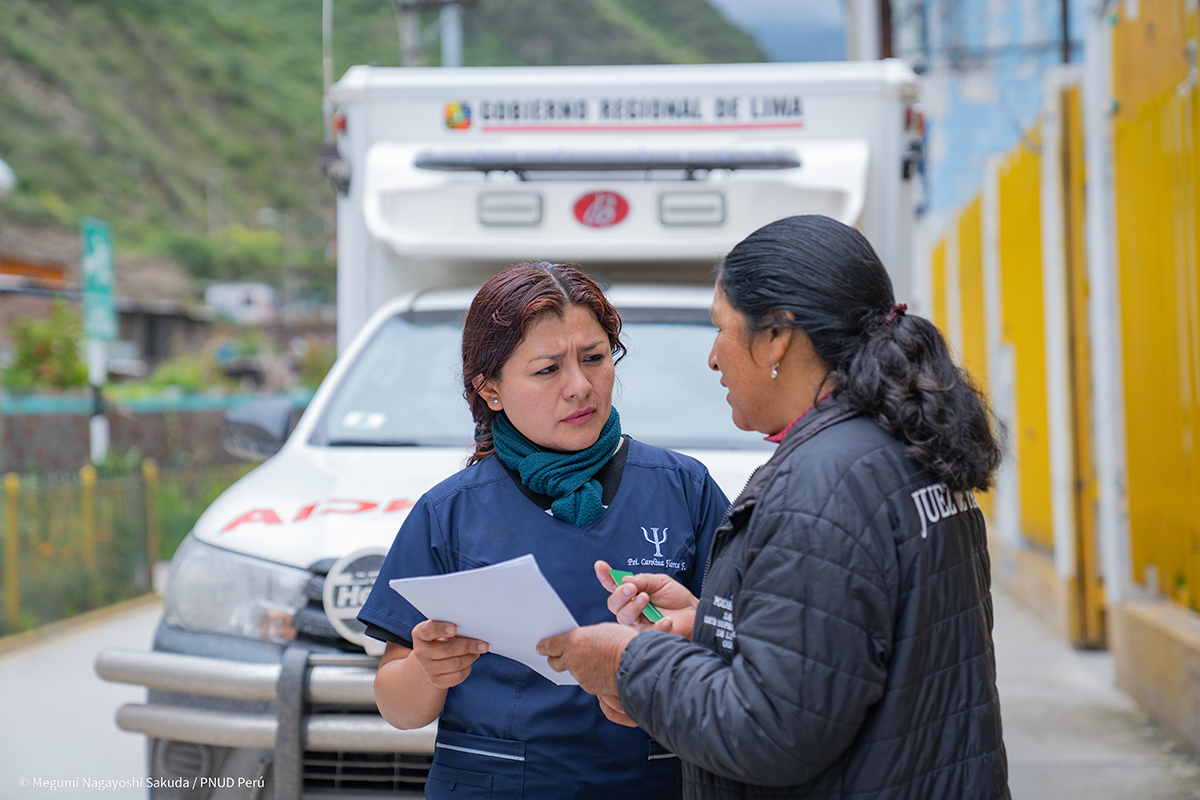 Two women discuss documents outside an ambulance in a rural area.