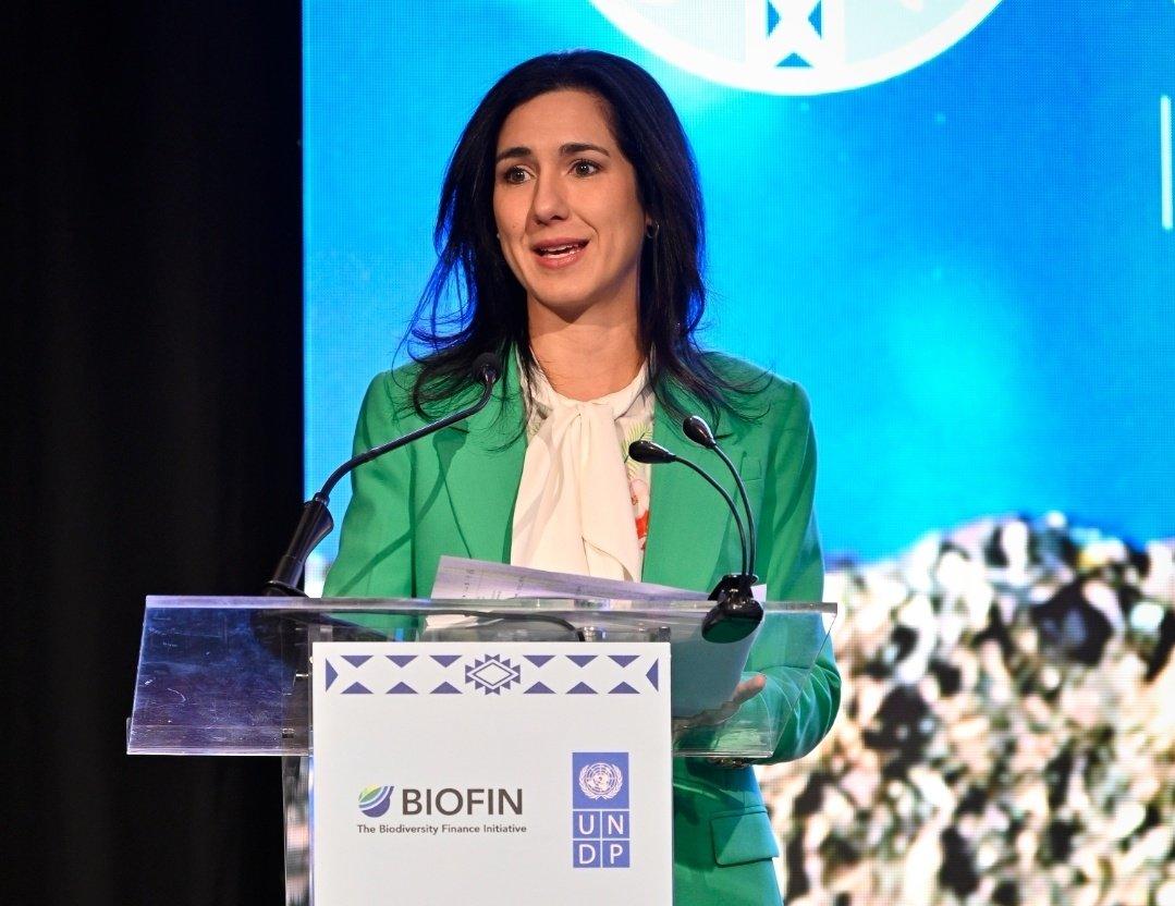A woman with dark hair wearing a green blazer speaks at a podium during an event.