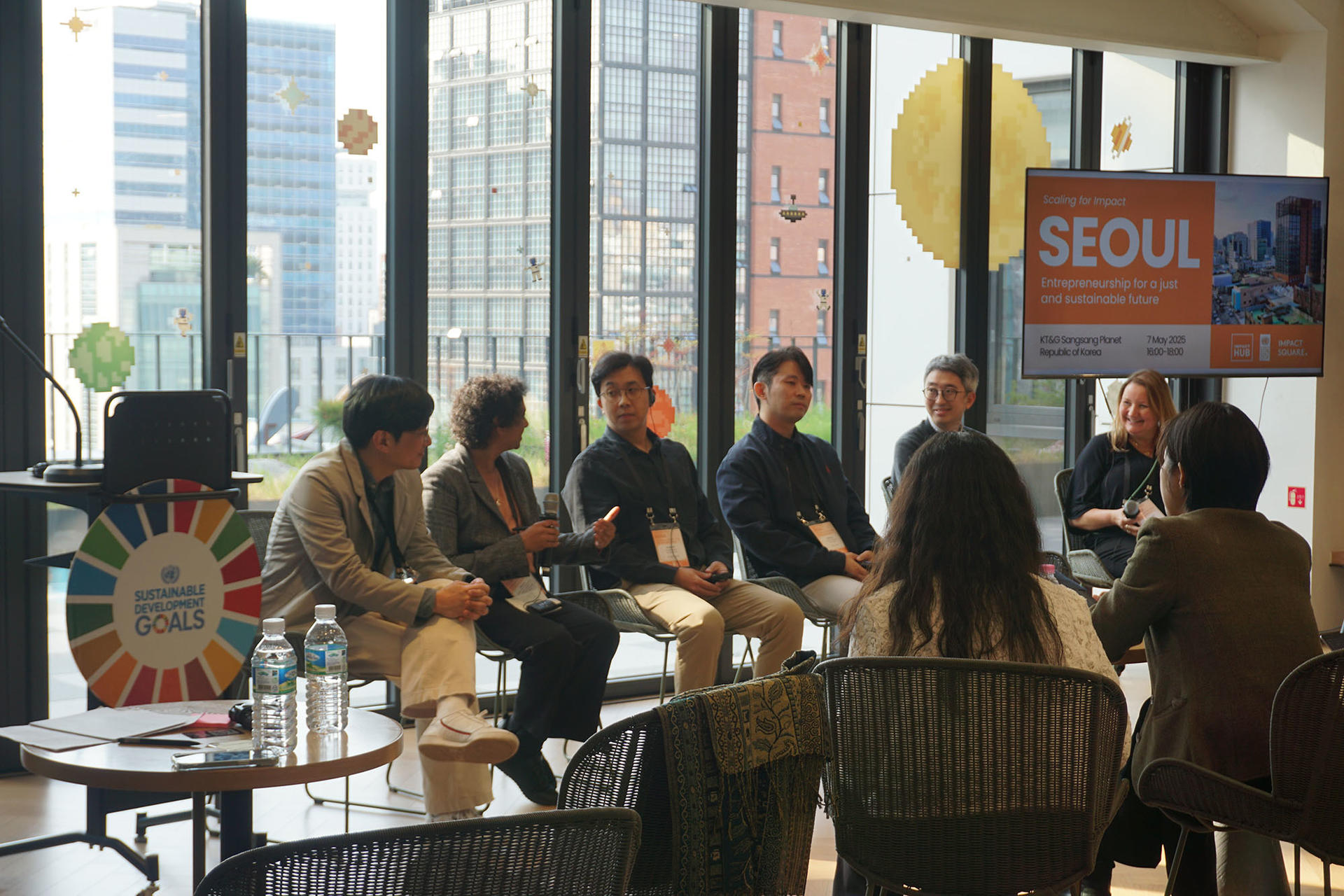 Five panelists seated at a table discussing in a brightly lit room with large windows.
