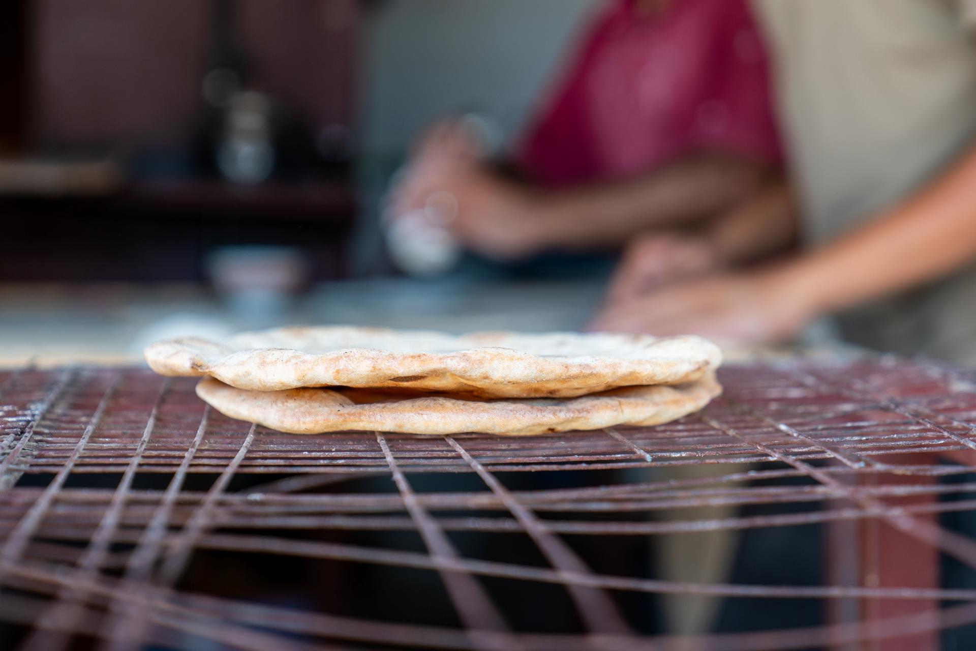 Two freshly made flatbreads stacked on a wooden surface, with hands preparing dough in the background.