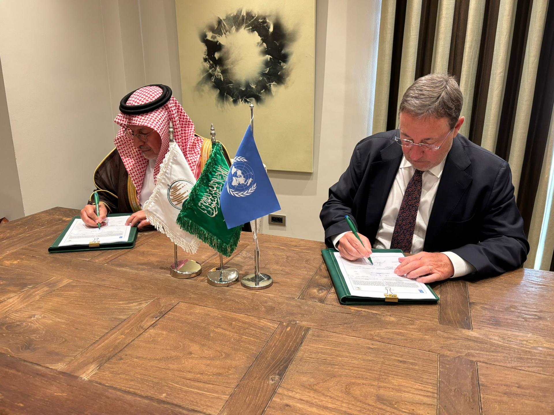 Two men, one in traditional Arab dress and one in formal attire, sign documents at a wooden table.
