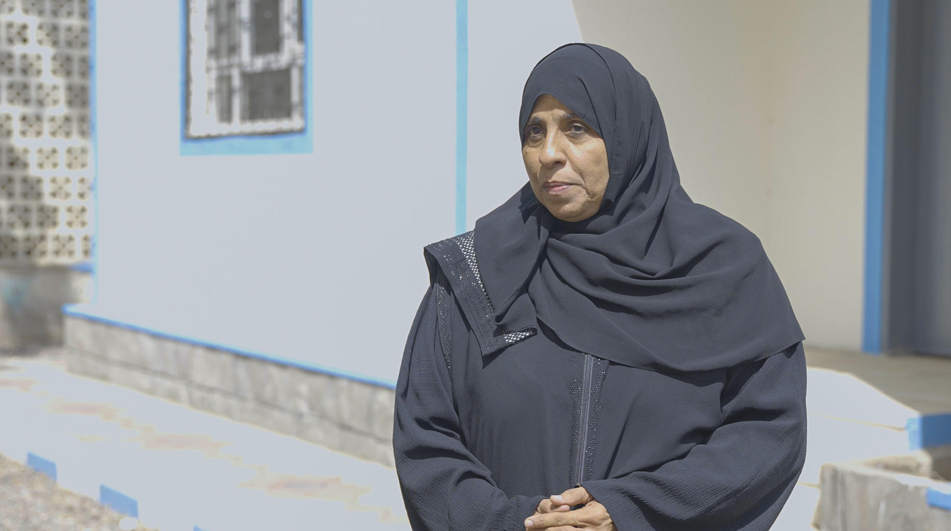 A woman in a black hijab stands outside a building, looking thoughtfully to the side.