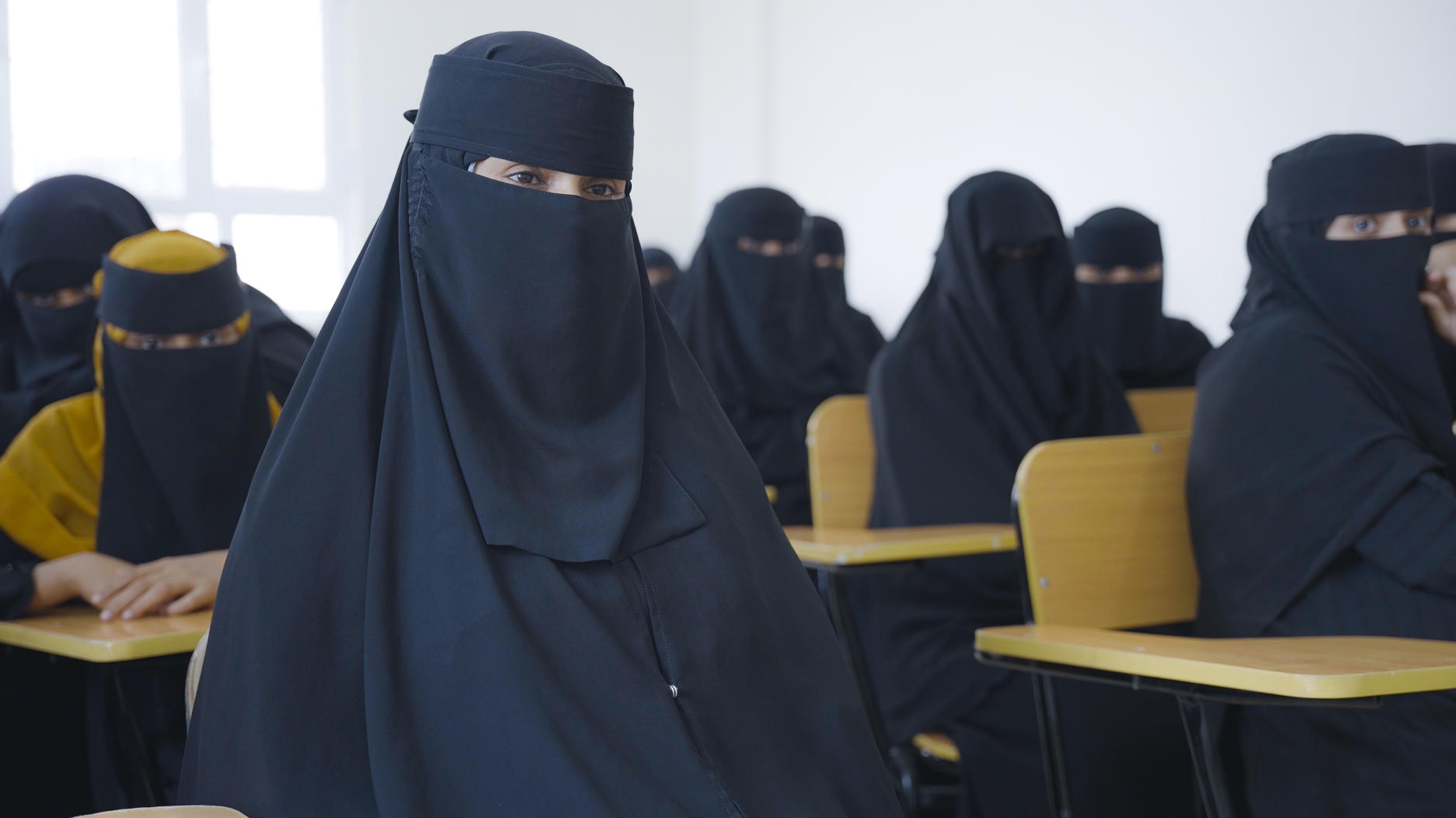 A classroom scene with women in black niqabs sitting at desks, focused and attentive.