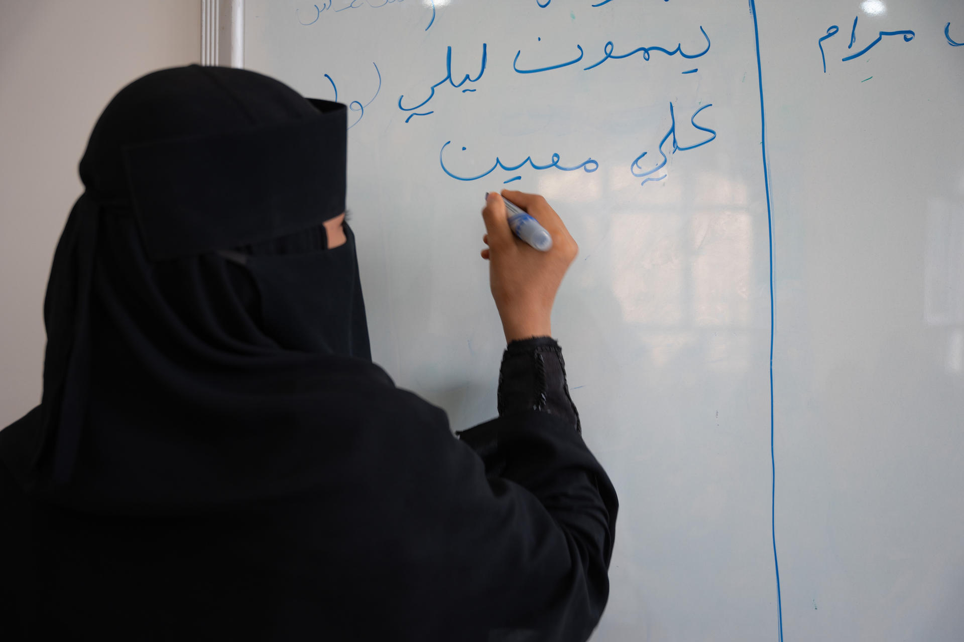 A woman in a black niqab writes on a whiteboard with a blue marker.