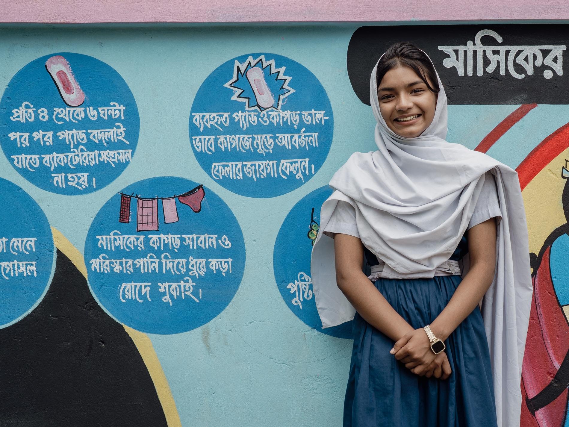 Young woman in a hijab smiles in front of a colorful educational wall mural.