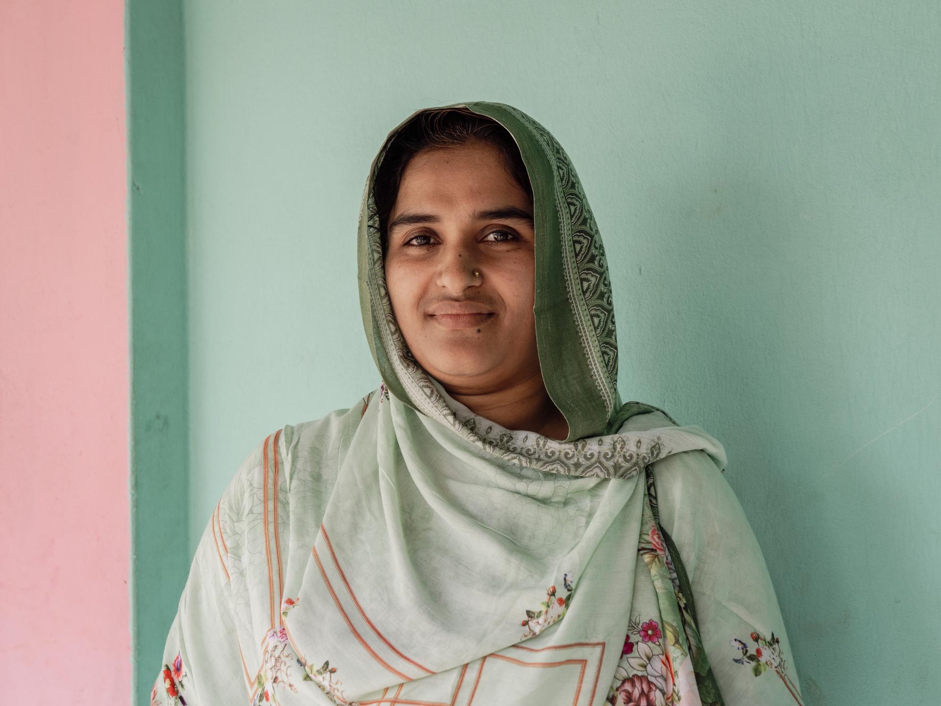 Smiling woman in a light green headscarf, standing against a pastel wall.