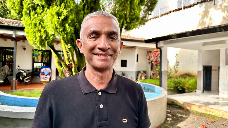 Smiling man in a black polo shirt stands outdoors in a garden setting.