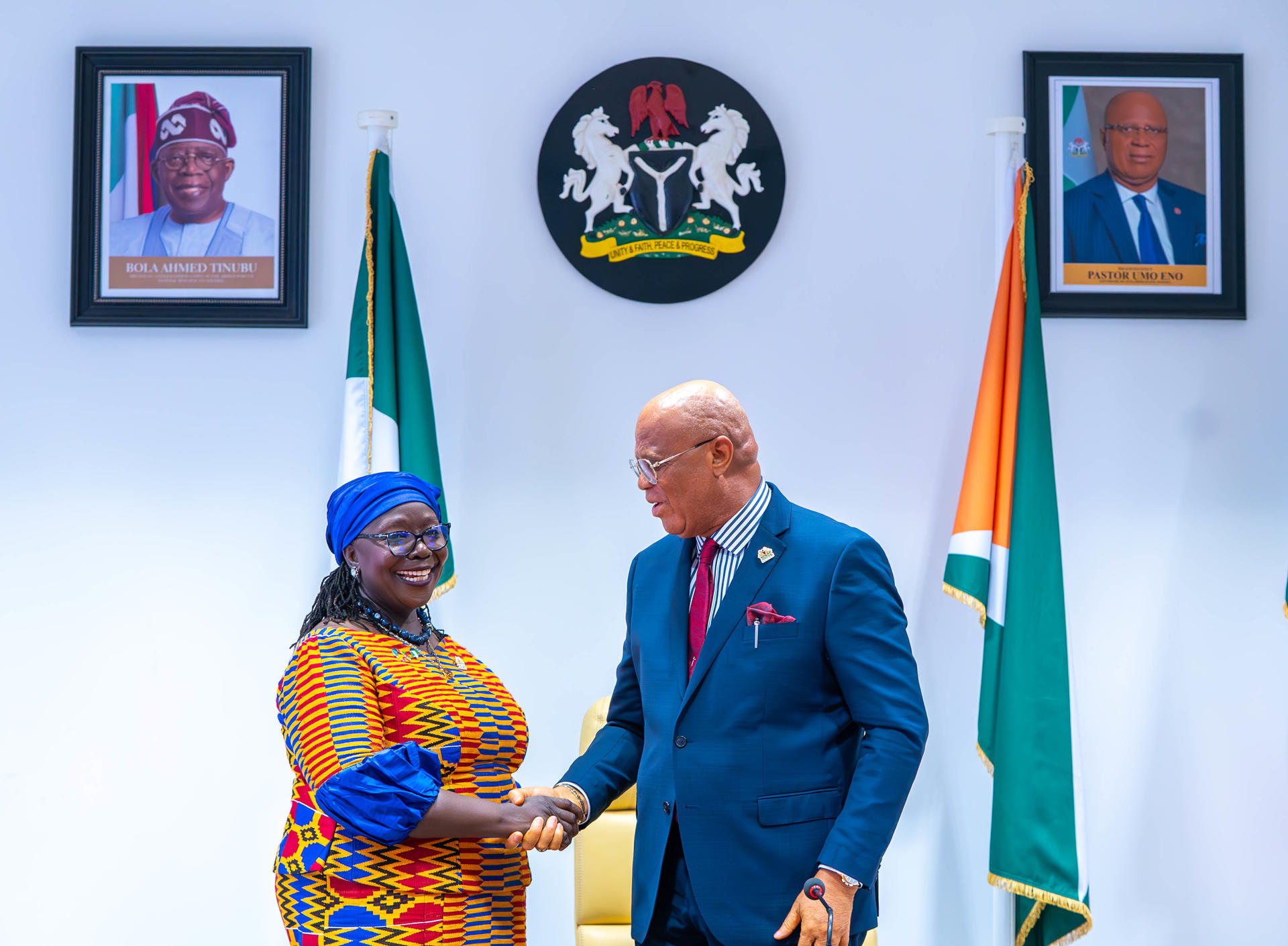 Two officials shake hands, smiling, with flags and portraits in the background.