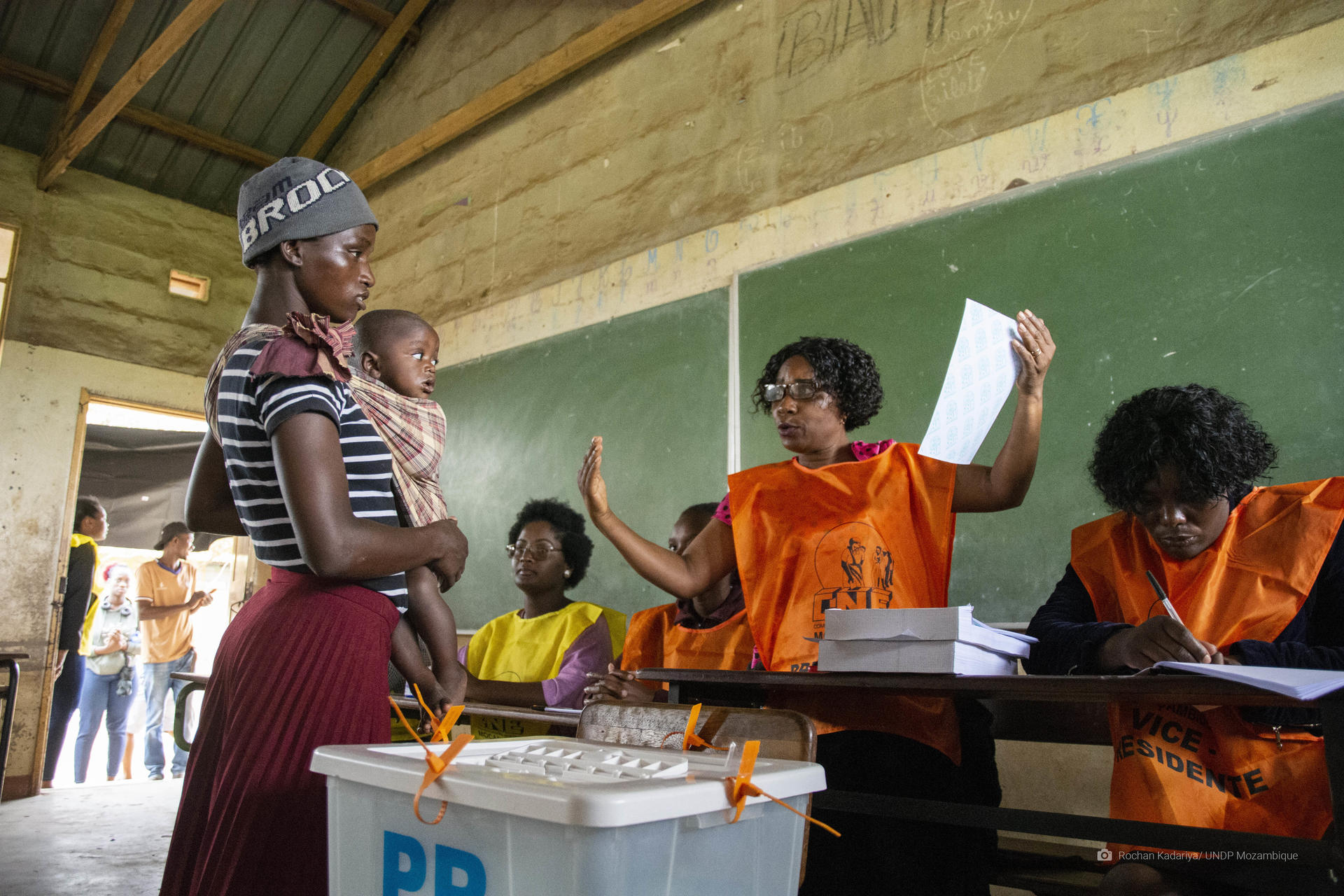 A woman holding a baby stands as she is given instructions on how to cast the ballots at a voting station.