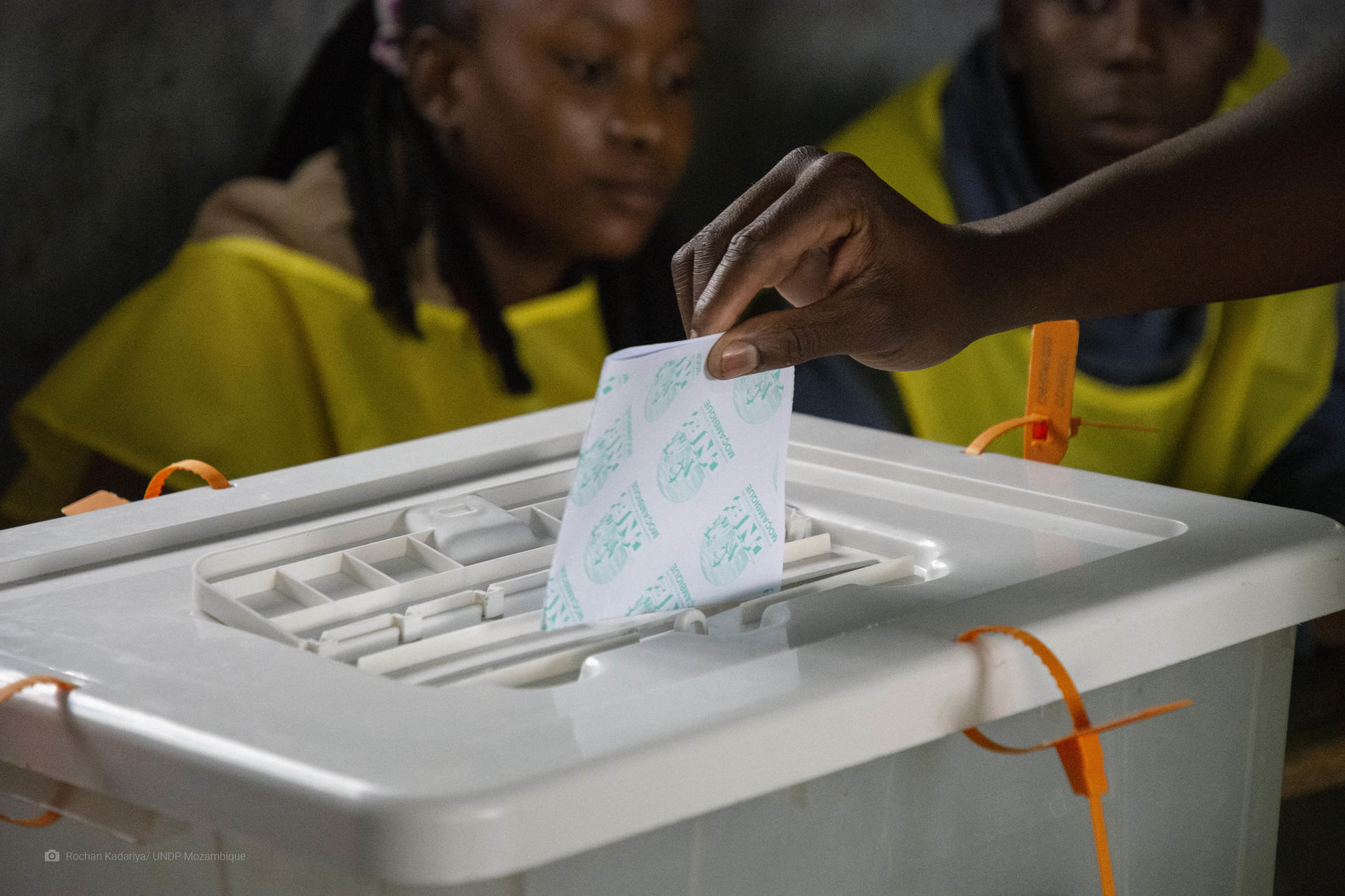 A hand dropping a ballot into a white ballot box, with two elections officers observing in the background.