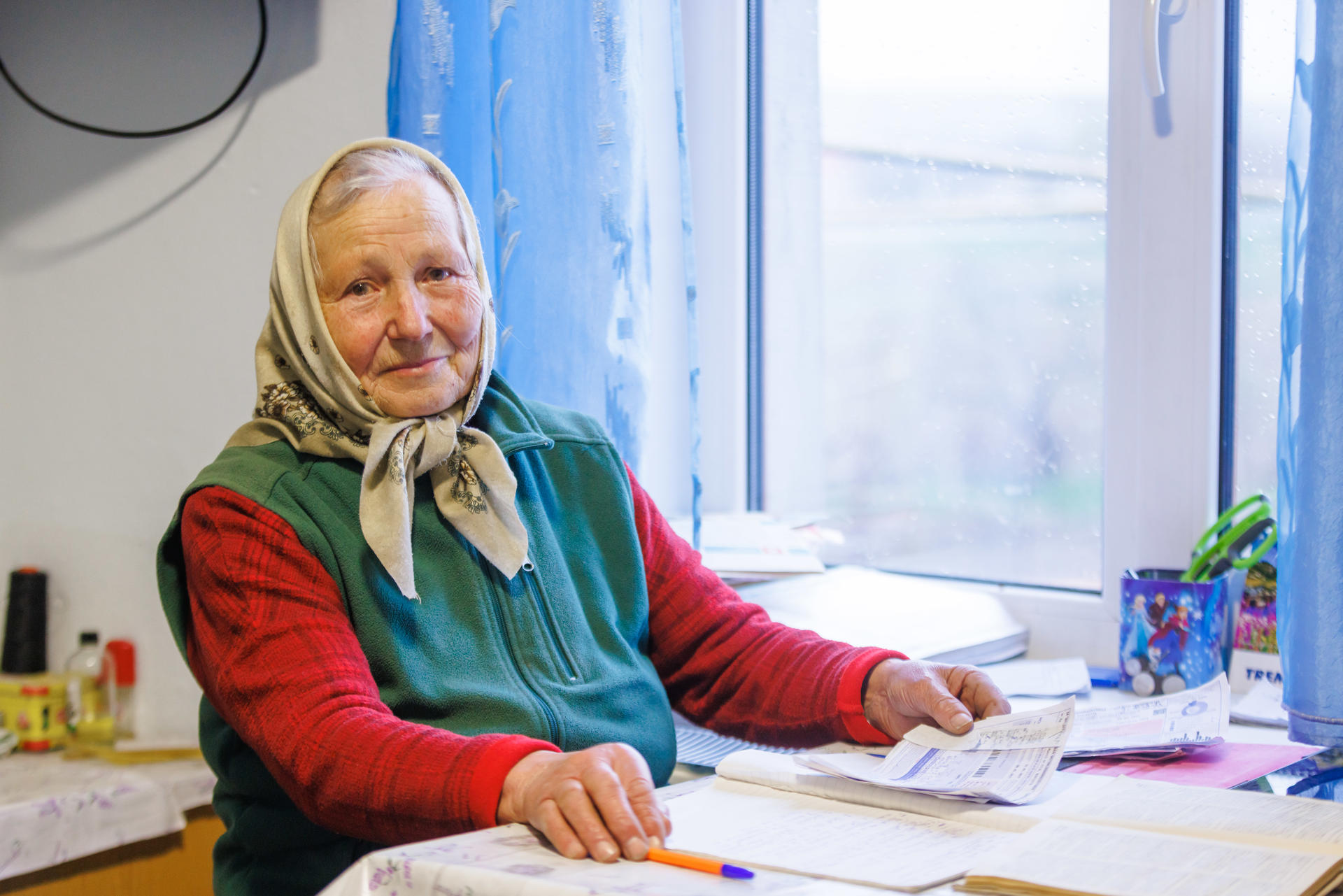Elderly woman with a headscarf smiling at a table by a window, surrounded by papers.