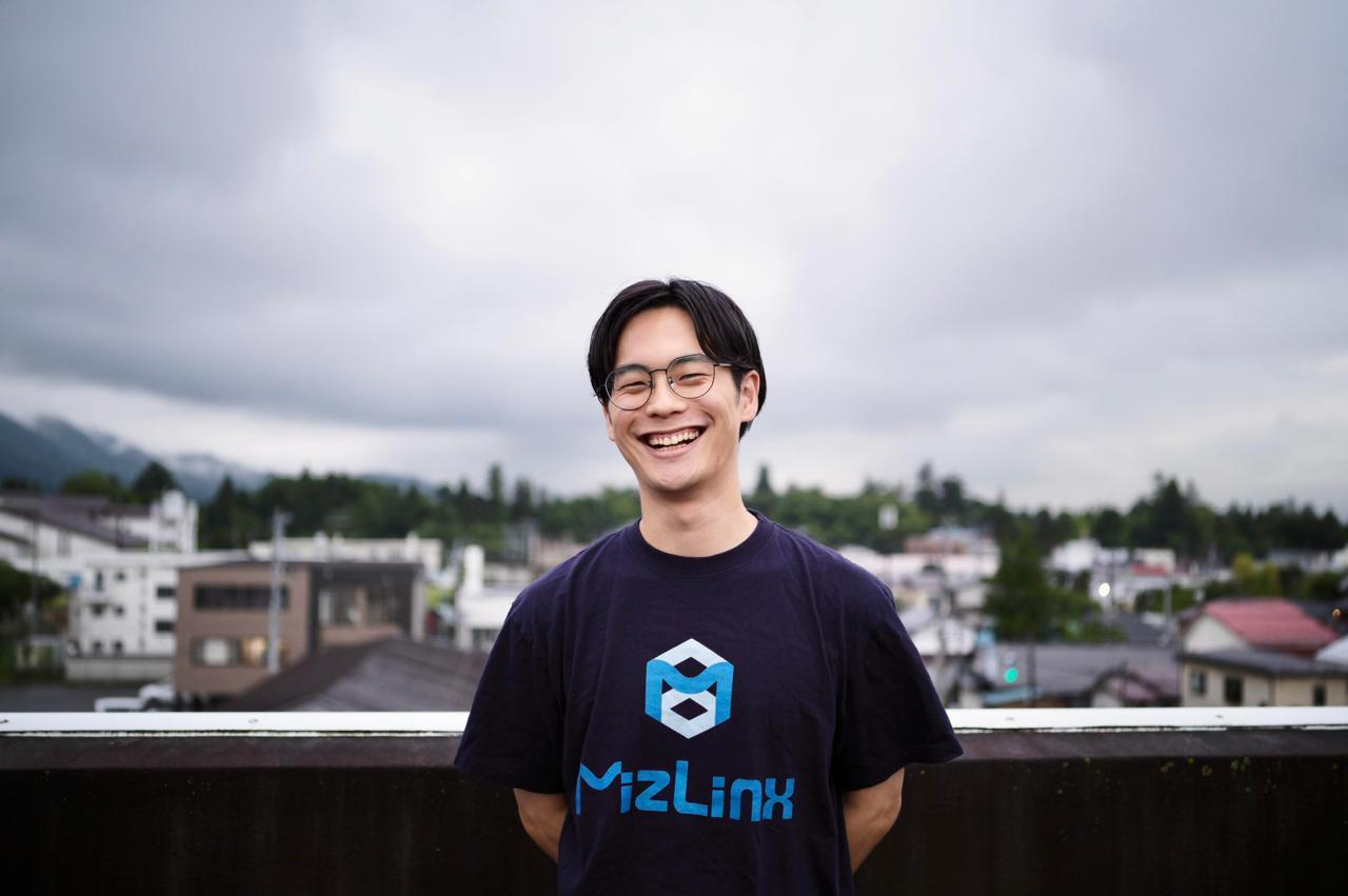 Smiling man in a black t-shirt stands outdoors with cloudy skies and buildings in the background.