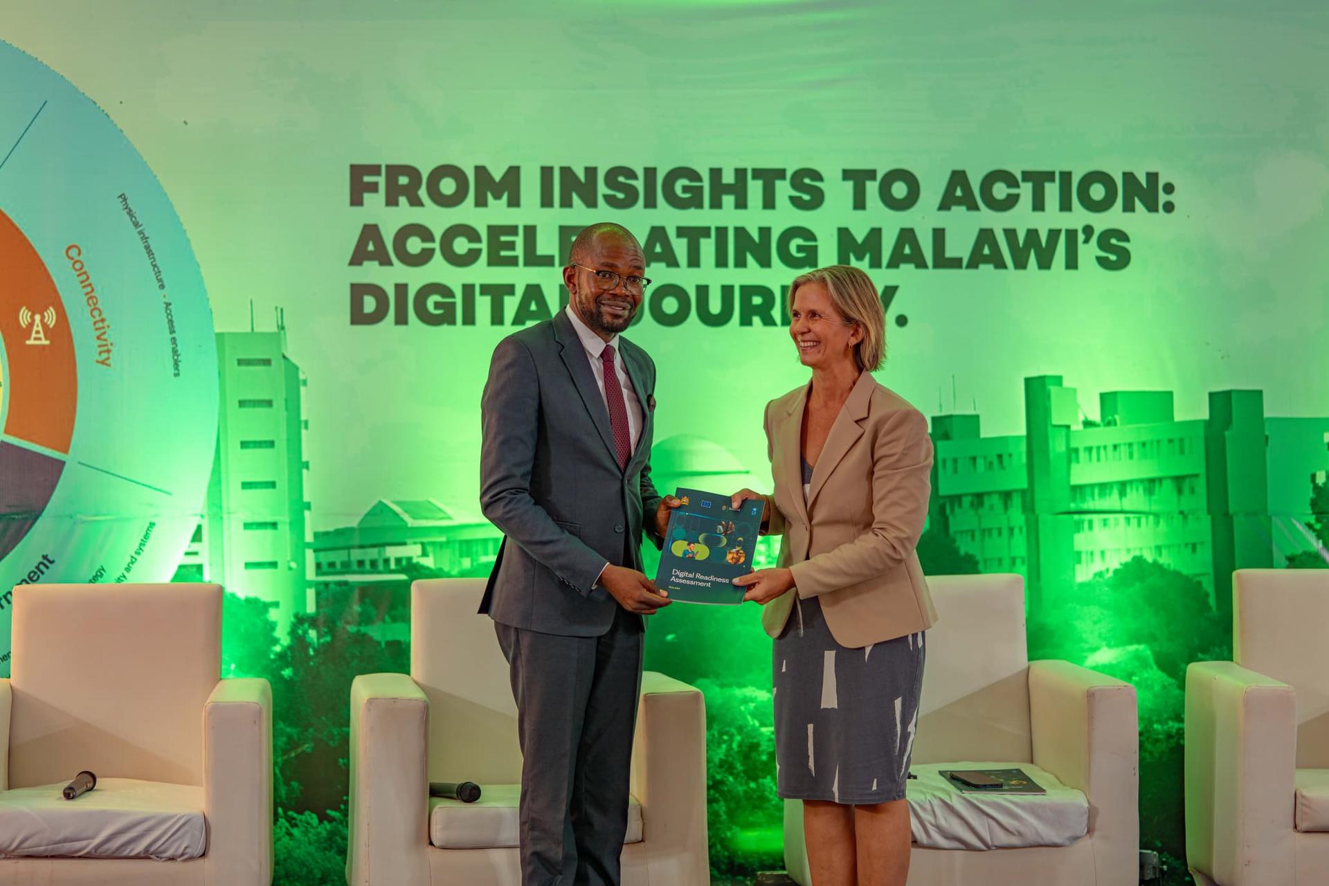 Two people exchanging a book on stage, with a backdrop featuring green lighting and text.
