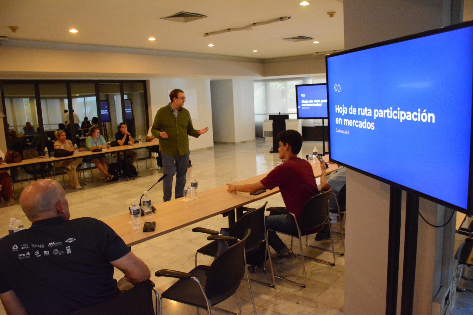 A speaker presents to an audience in a conference room, with a screen displaying text.