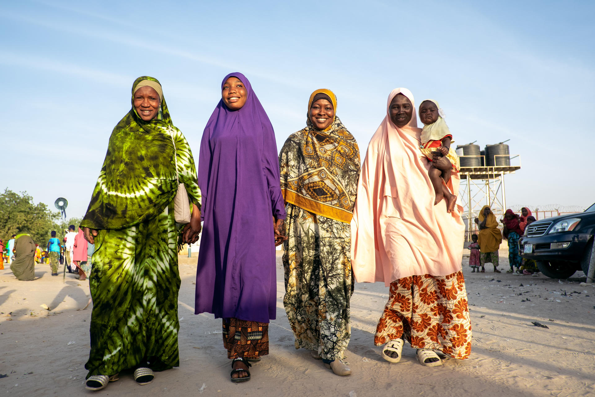Four women in colorful traditional attire walk together, smiling in a sunny outdoor setting.
