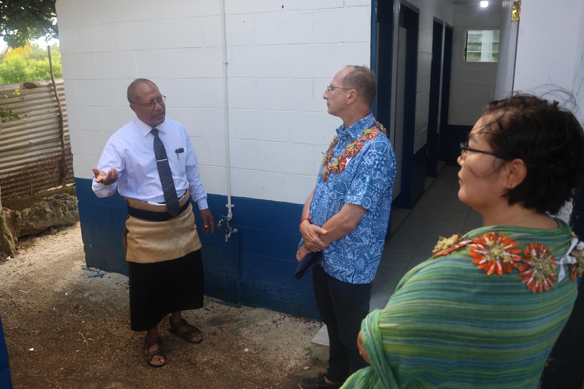 Three individuals converse outside a white and blue building, with one gesturing while speaking.